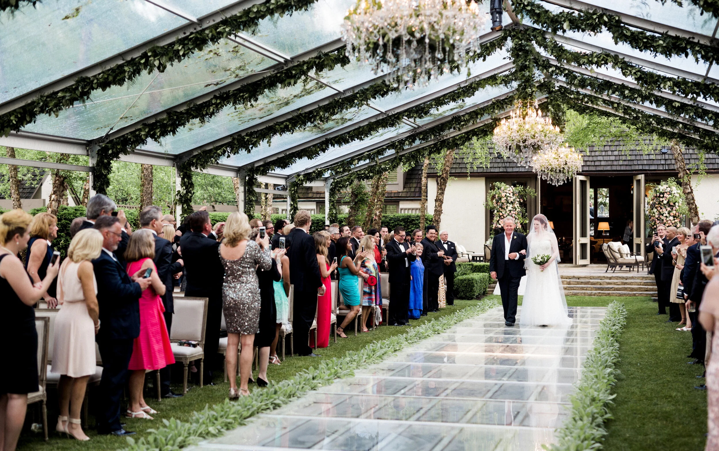 Wedding ceremony under a clear tent with guests standing, a bride walking down the aisle with an escort, surrounded by greenery and chandeliers.