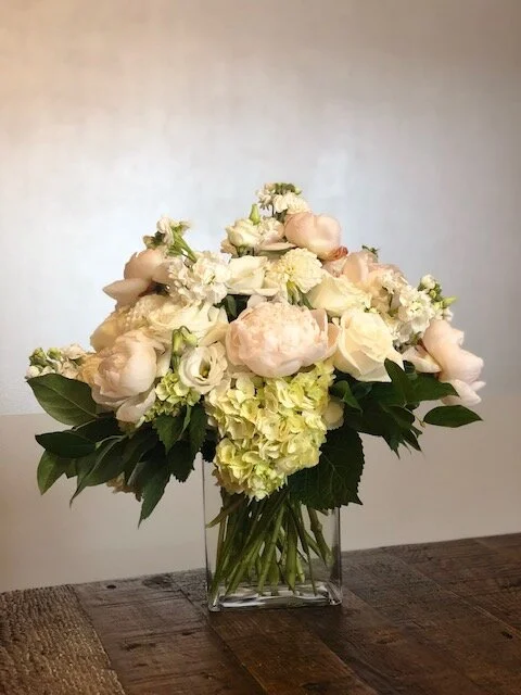 A bouquet of pastel-colored peonies, roses, hydrangeas, and greenery in a clear glass vase on a wooden table.