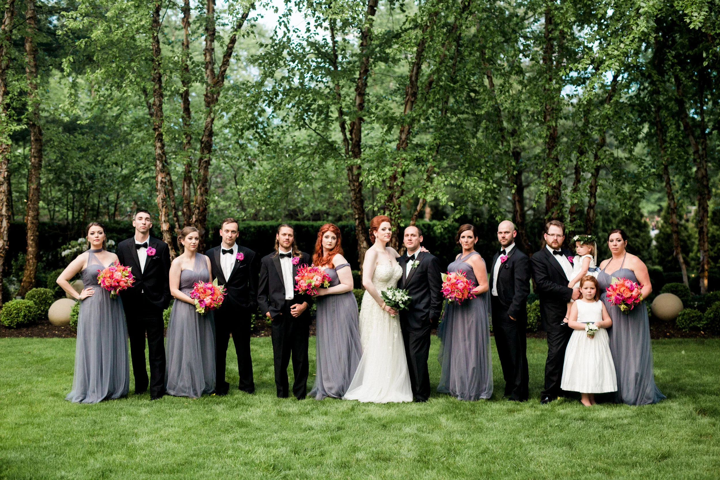 Wedding party posing outdoors on grass, with bridesmaids in gray dresses holding pink and orange flowers, groomsmen in black suits, and a bride and groom in the center.
