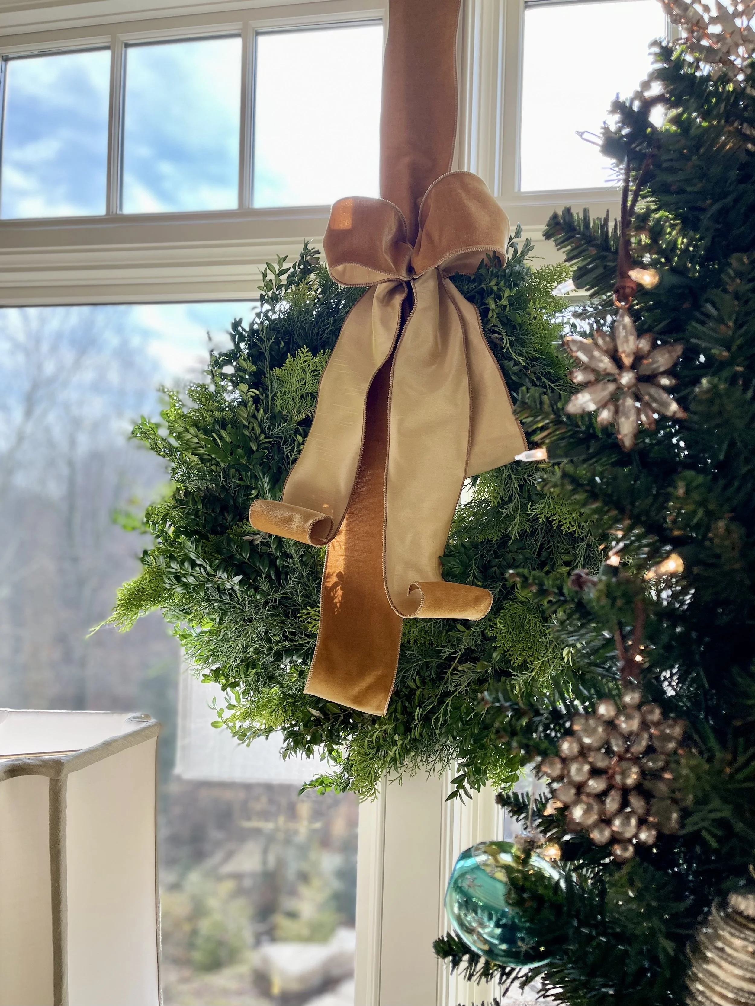 Close-up of a festive holiday wreath hanging on a window, decorated with a large gold and brown velvet ribbon, surrounded by Christmas tree branches with ornaments, in a bright room with sunlight streaming in.