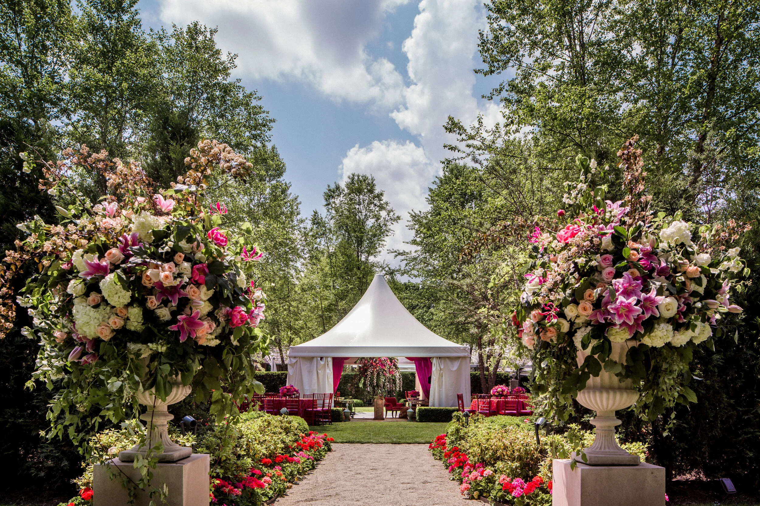 Outdoor wedding event with a white tent surrounded by lush greenery, large floral arrangements with pink and white flowers, red chairs, and a path lined with flowers leading to the tent.