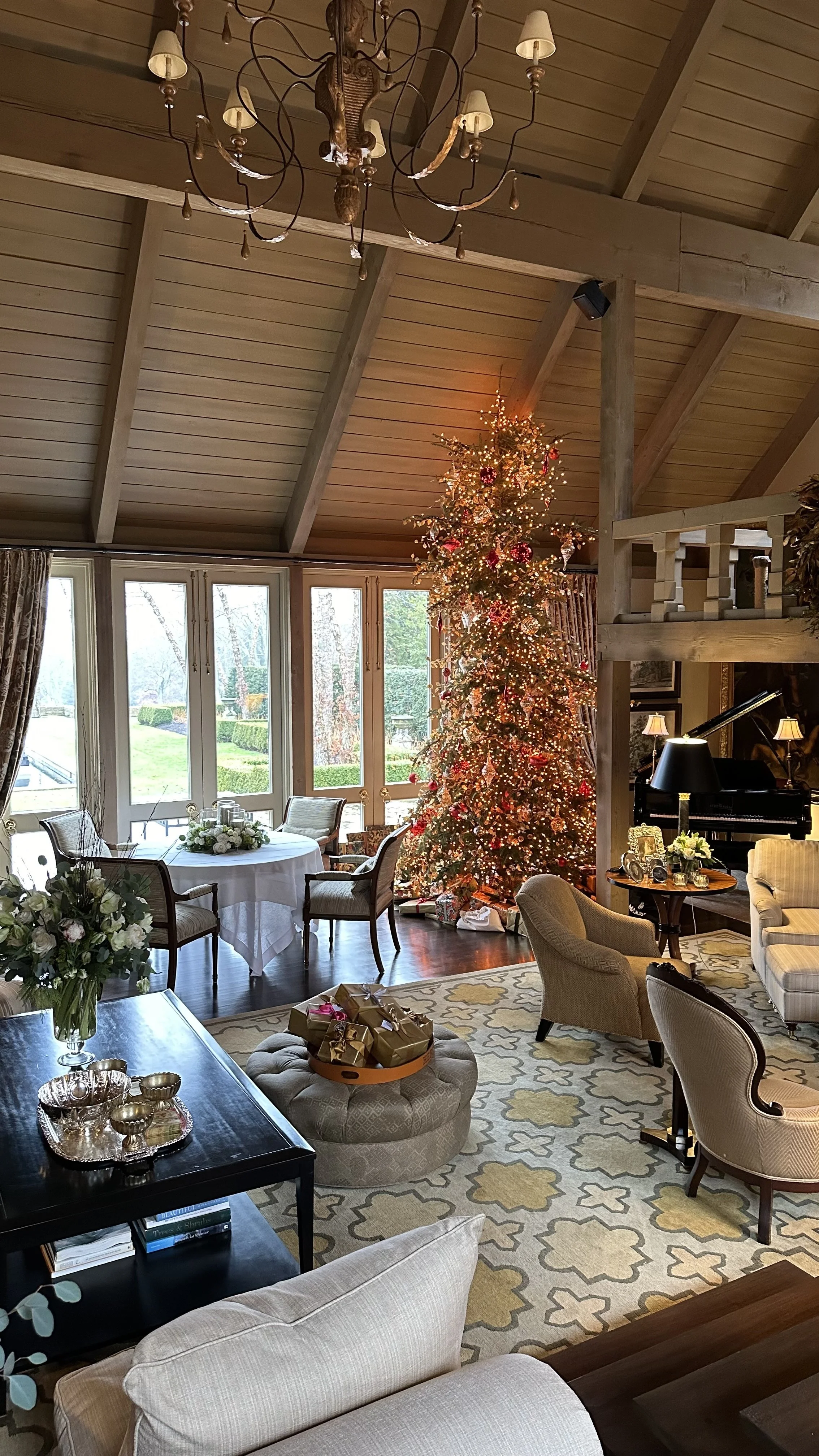 Living room decorated for Christmas with a large illuminated Christmas tree near the window, a chandelier hanging from the ceiling, a round table with chairs, a piano, and wrapped gifts on an ottoman.