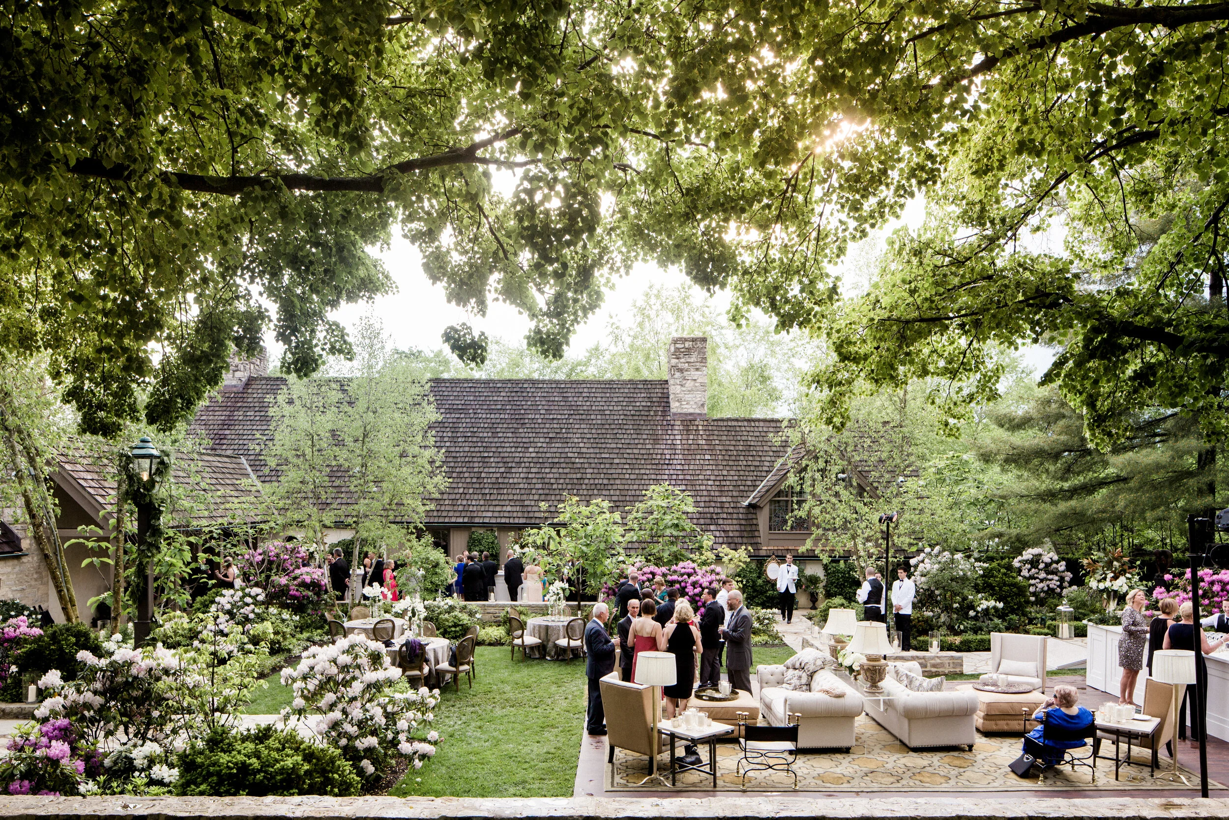 Outdoor garden party with guests mingling, surrounded by flowers and greenery, with tables and outdoor seating arrangements.
