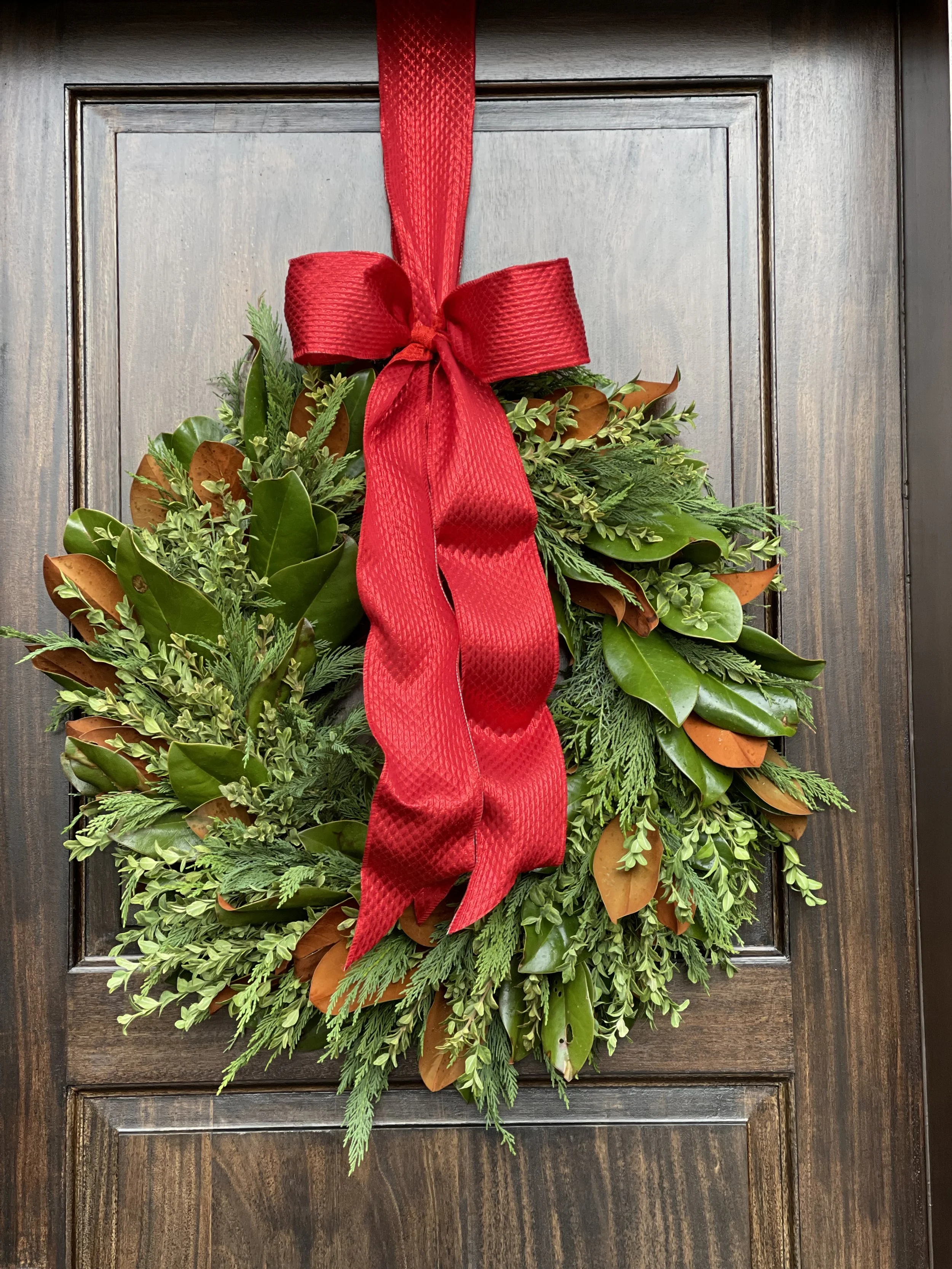 A Christmas wreath with green foliage and brown accents, decorated with a large red ribbon bow, hanging on a wooden door.
