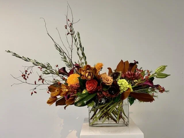 Floral arrangement with roses, lilies, hydrangeas, and assorted greenery in a glass vase on a white pedestal.