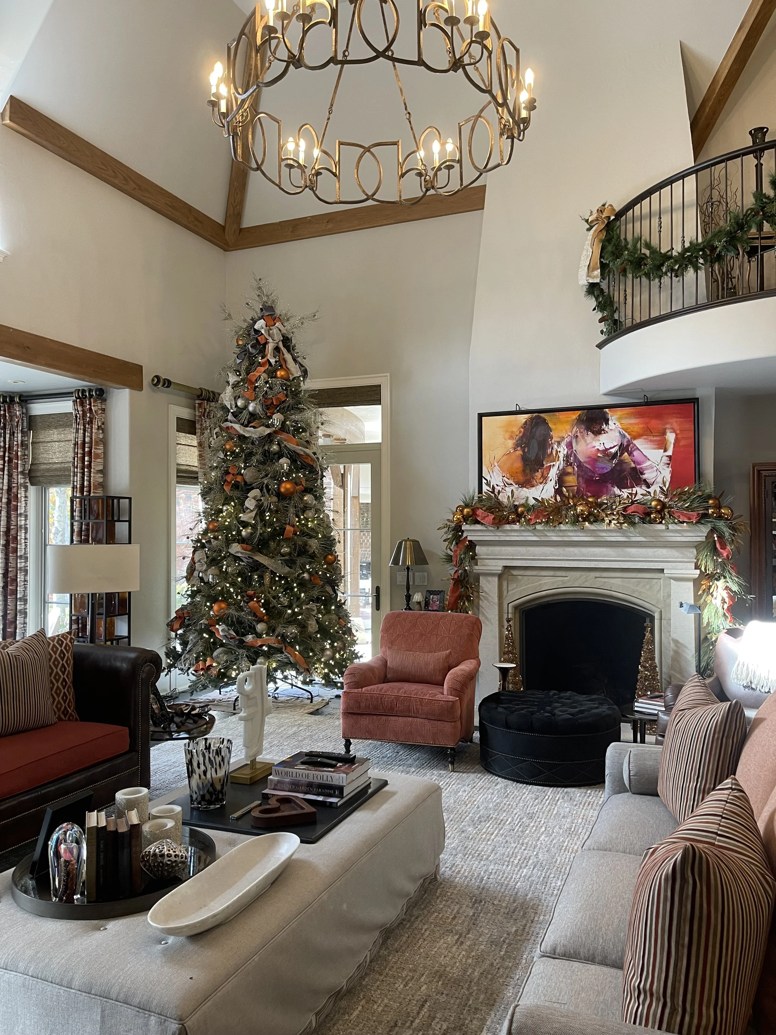 Living room decorated for Christmas with a large tree, a fireplace adorned with garland, and a couch and armchair, illuminated by a chandelier.