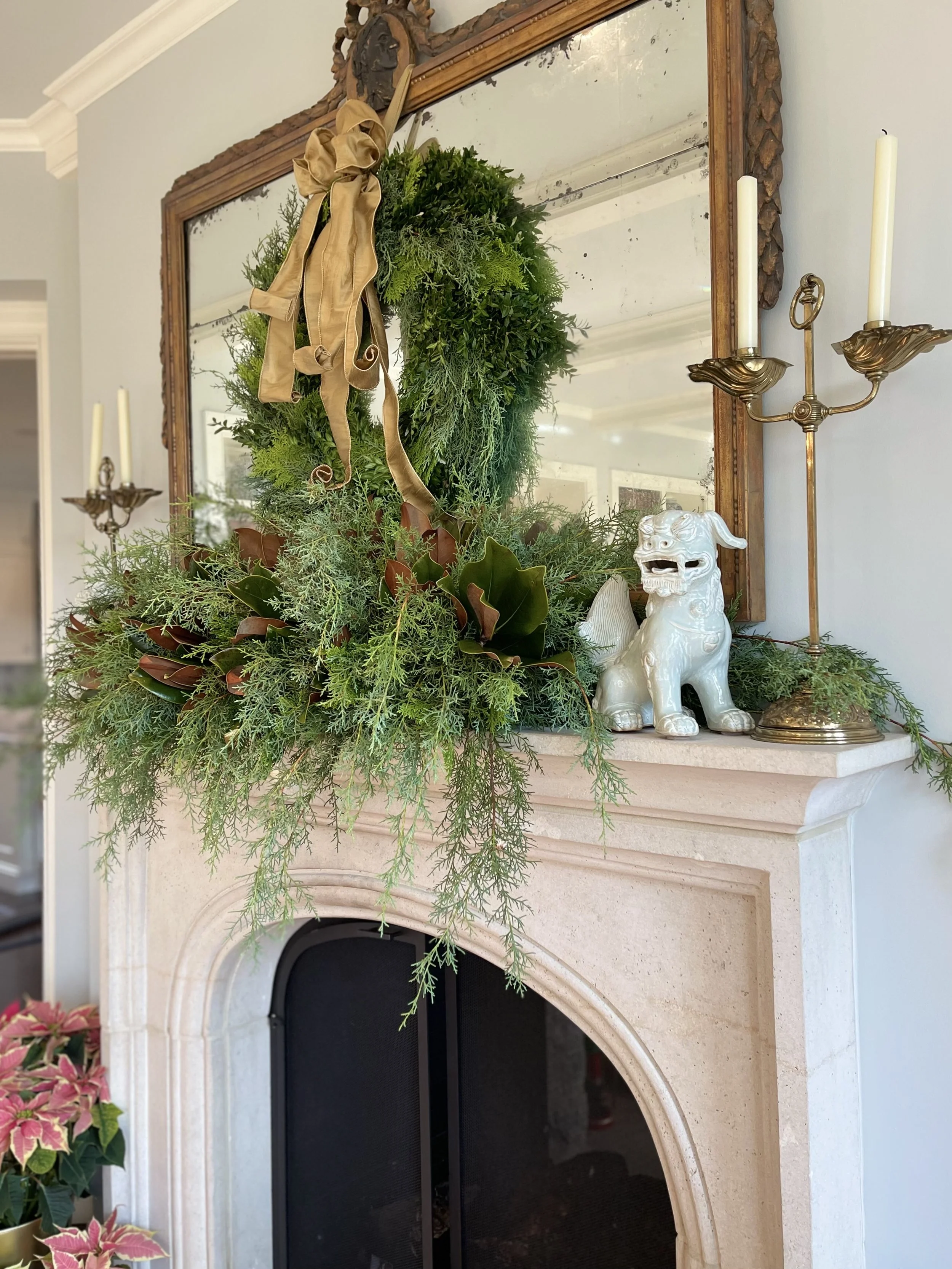 Decorative fireplace mantel with a green wreath, gold ribbon, a white Chinese guardian lion statue, a gold candle holder, and a mirror.