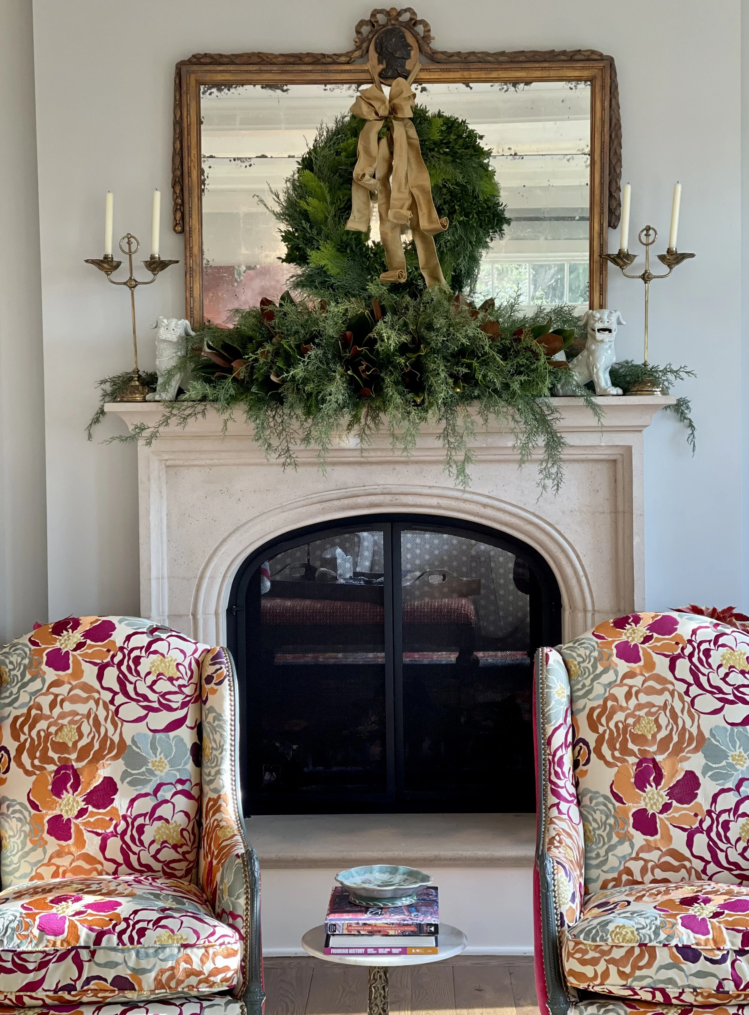 A living room fireplace decorated with greenery and a large wreath with a gold ribbon, framed by a large mirror, flanked by candelabras and ceramic dog figurines. Two colorful floral armchairs sit in front of the fireplace, with a small side table an