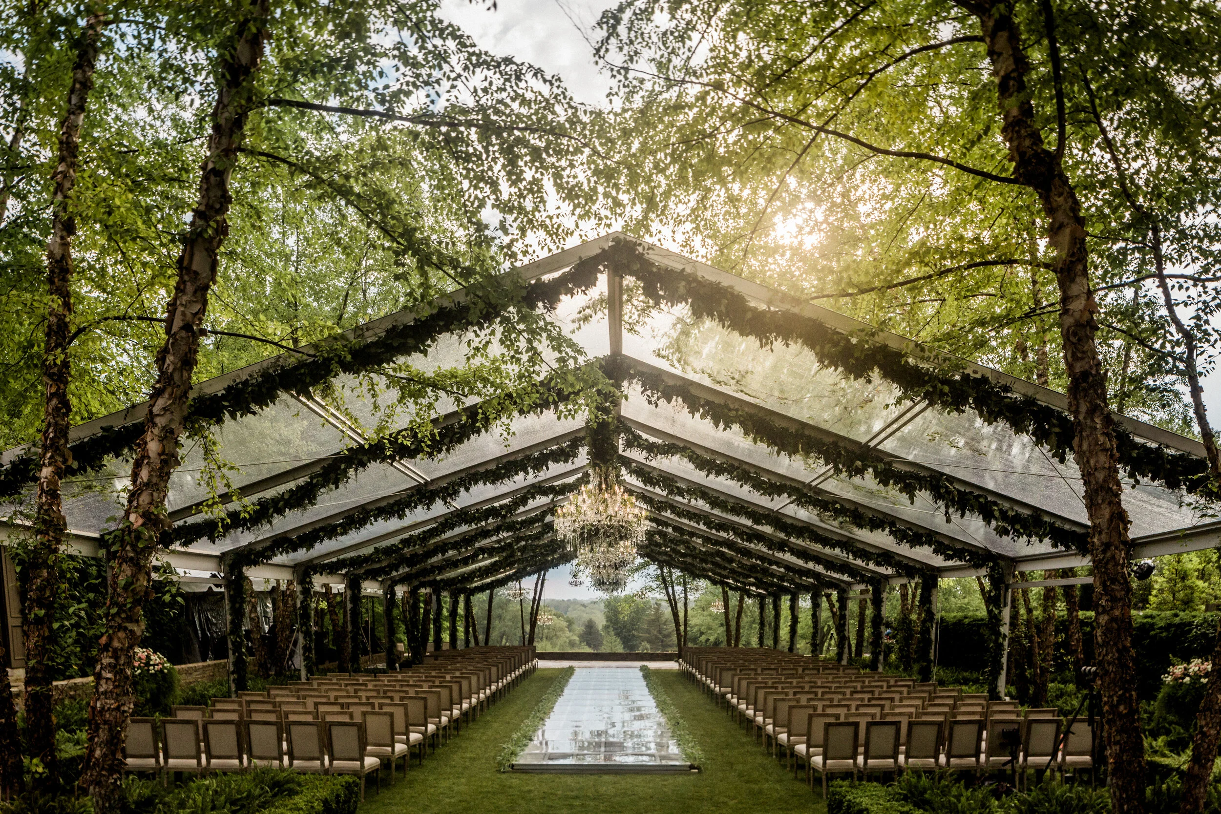 Outdoor wedding ceremony setup under a transparent tent with rows of chairs, surrounded by trees and greenery, featuring a reflective aisle and chandeliers.