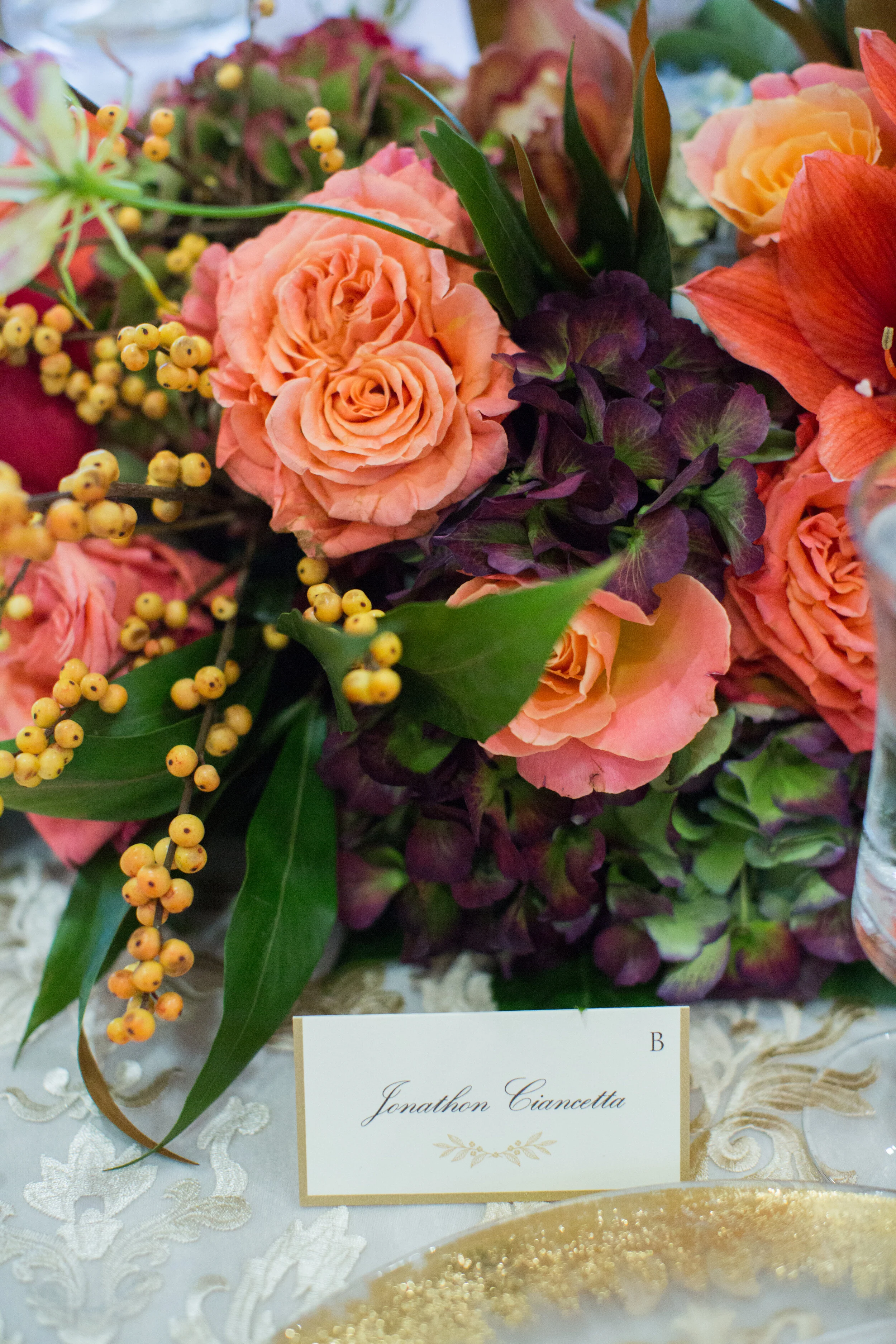 Floral table centerpiece with orange roses, purple hydrangeas, and yellow berries. Place card marked B with elegant script. Lace tablecloth and gold-rimmed plate visible.