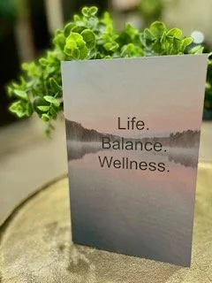 A booklet or card with a serene nature scene and the words 'Life. Balance. Wellness.' printed on it, placed among green plants.