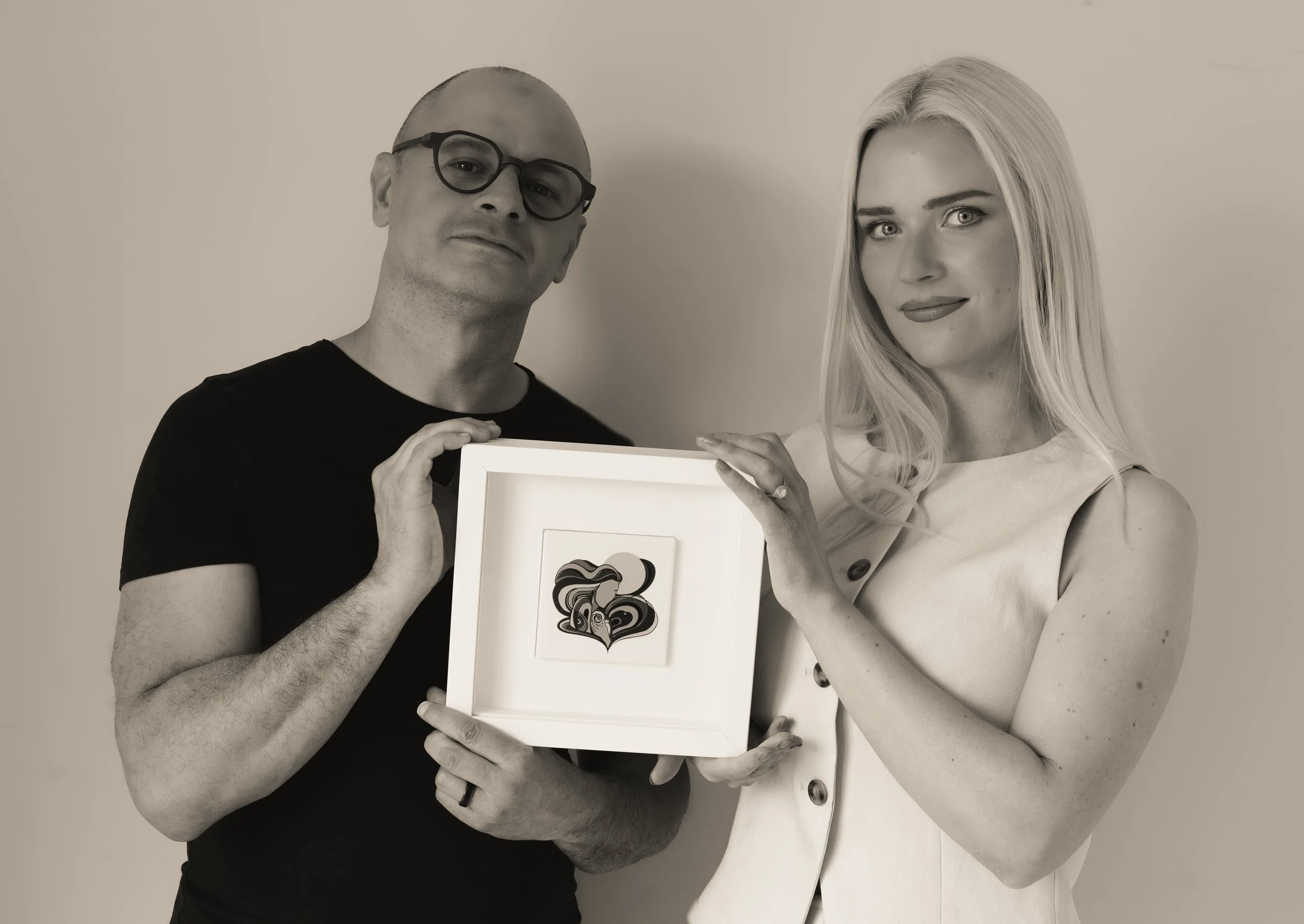A man with glasses and a woman with long blonde hair holding a framed abstract heart artwork together.