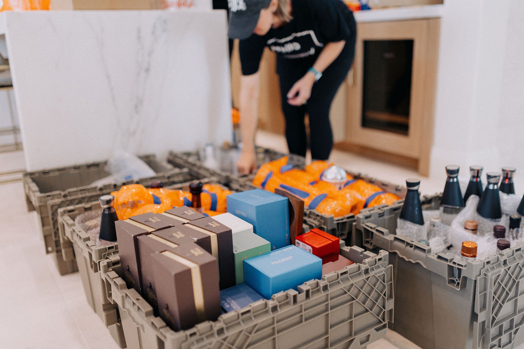 Person organizing crates of assorted bottled beverages and boxed items in a kitchen.