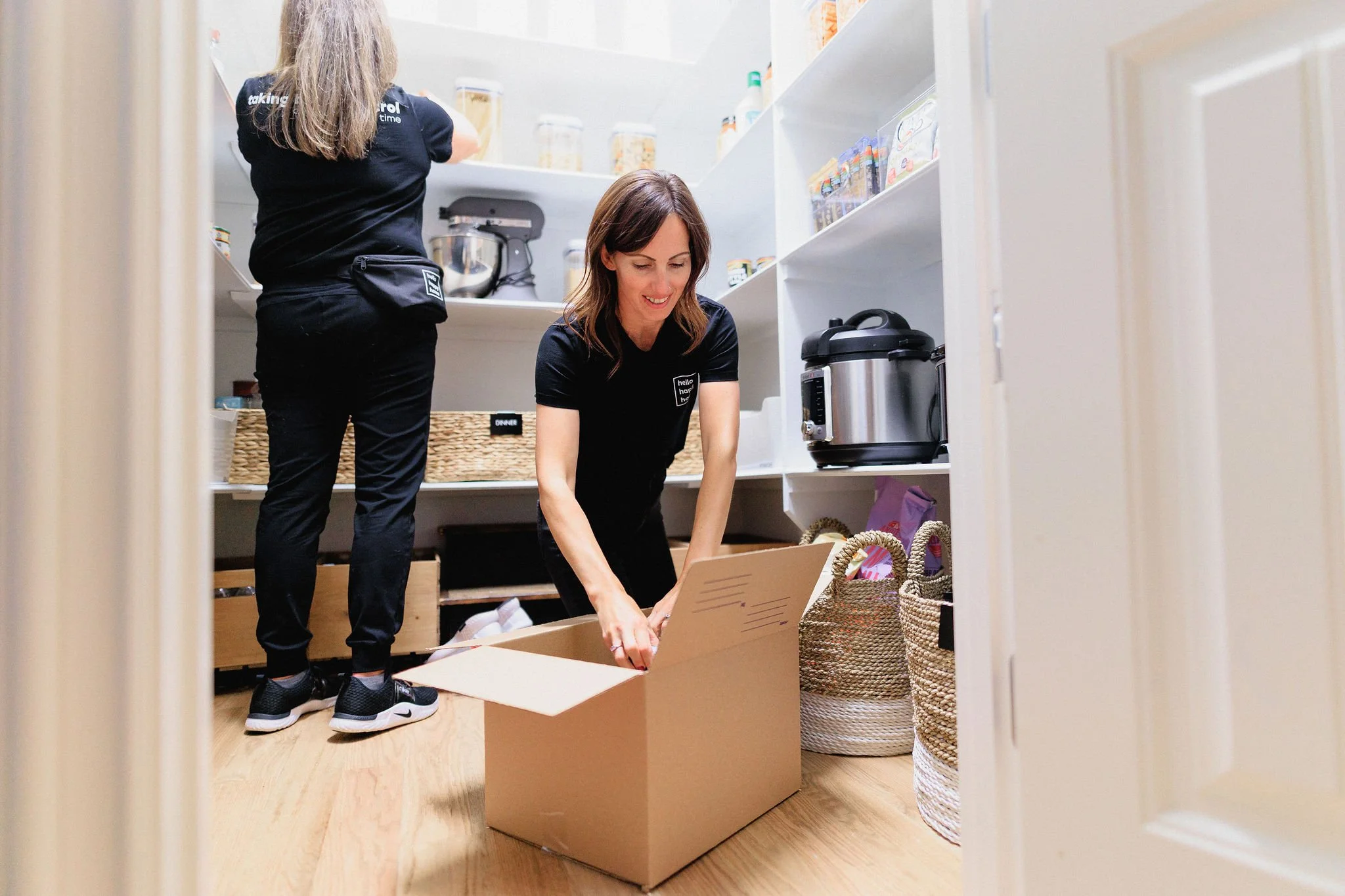 Two women unpacking food containers and organizing a pantry with baskets and kitchen appliances.