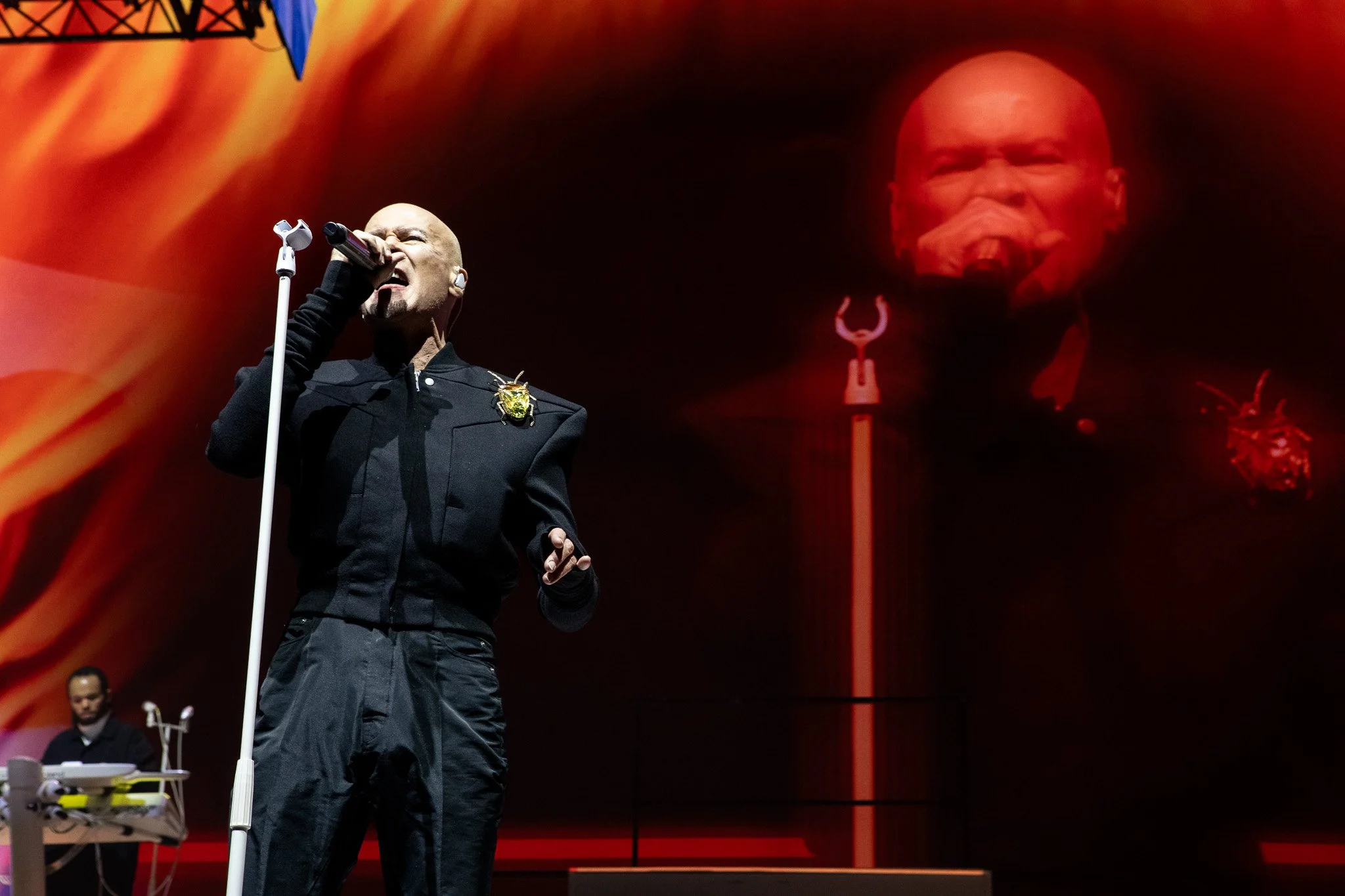 The Human League singer Phil Oakey in a black suit singing into a microphone stand. The video design in the background is red, by STUDIO/