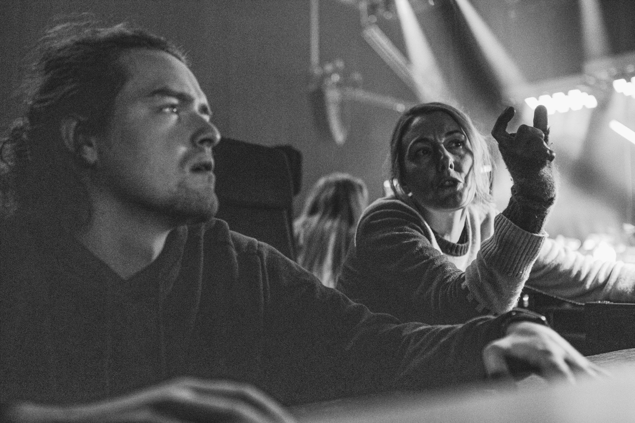 Black and white photo of two people talking in a rehearsal venue. The man on the left has long hair and is looking ahead, while the woman on the right is gesturing with her hand and talking. She is STUDIO/ editor, Emma.