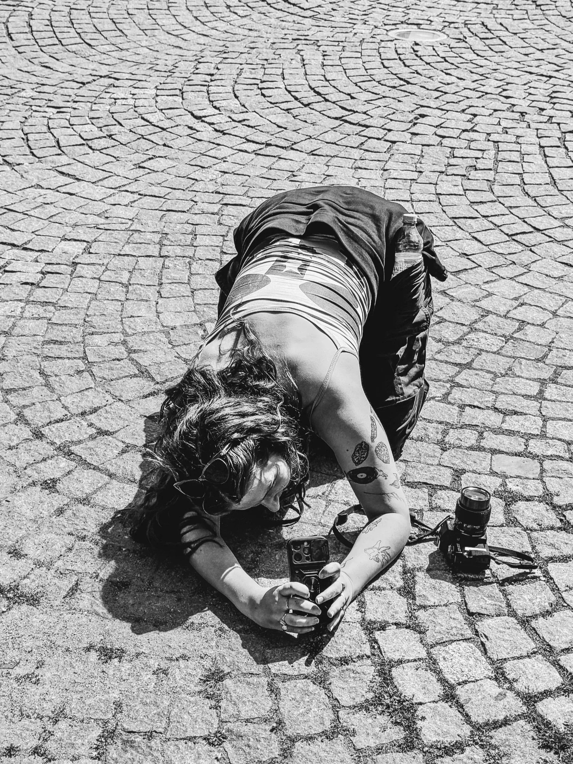 A woman is lying on a cobblestone street, taking a photo with her smartphone. She has photography equipment around her. She is STUDIO/ social media manager and photographer, Rosie.