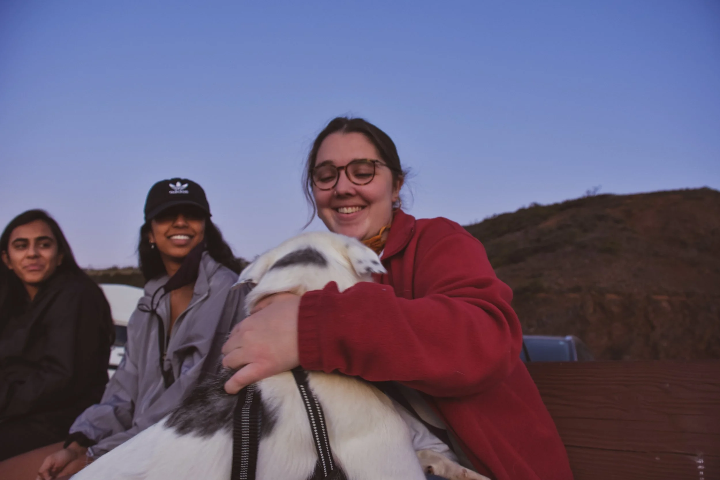 A group of young women and a dog outside during sunset with a hill in the background.