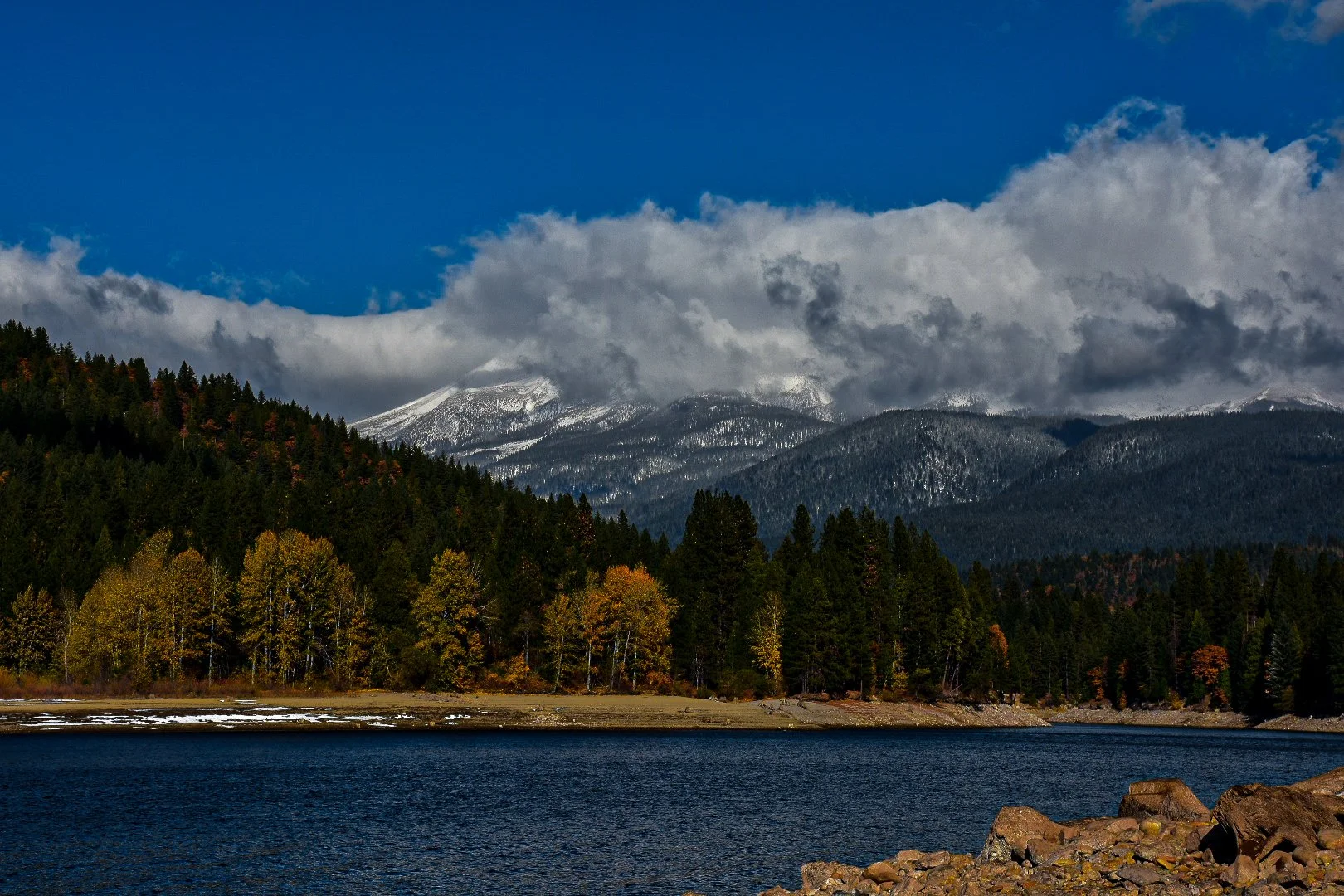 Mountain landscape with snow-capped peaks, a forested hillside, and a lake in the foreground.