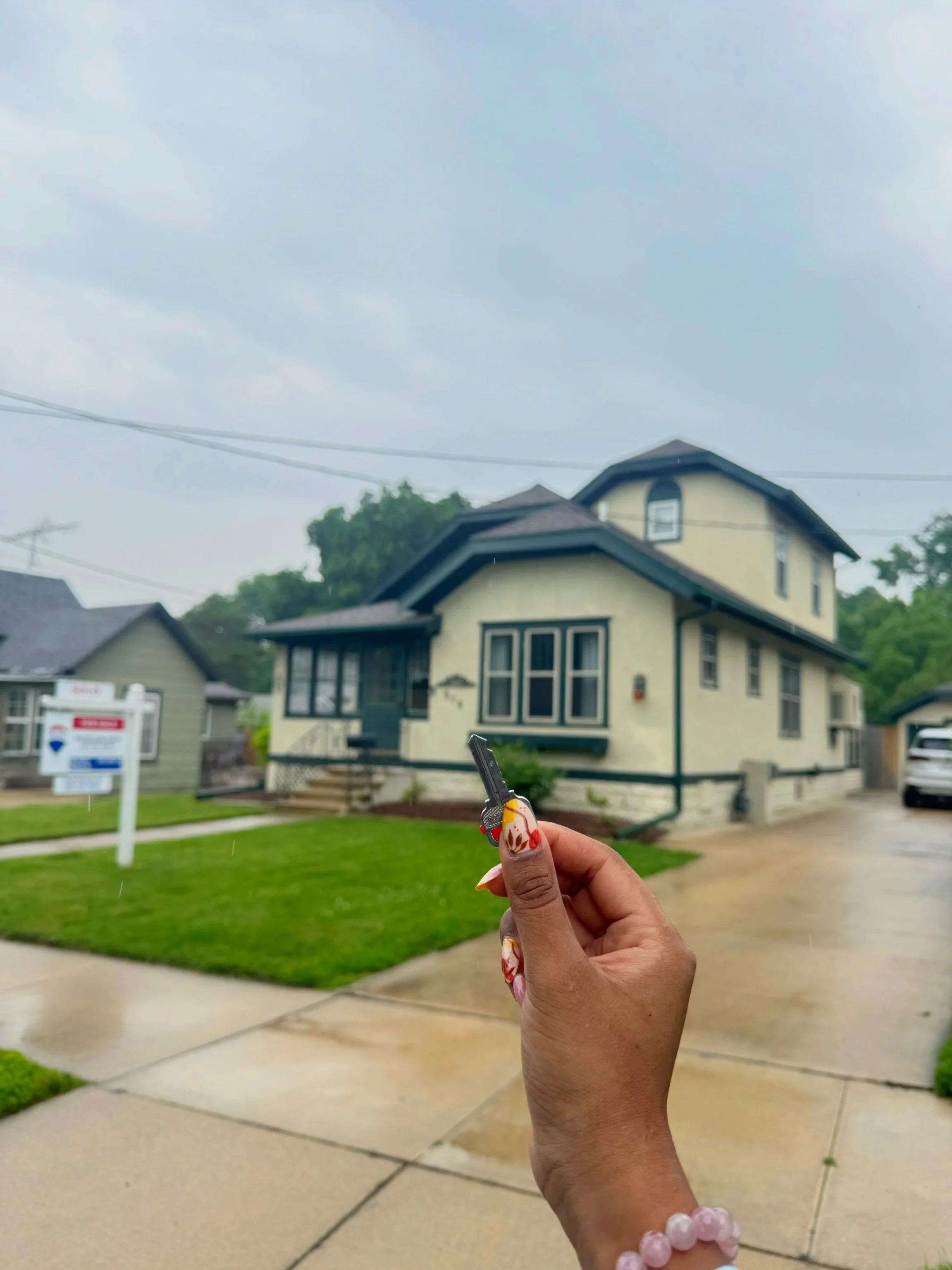 Person holding a set of house keys in front of a yellow house with black trim, on a rainy day.