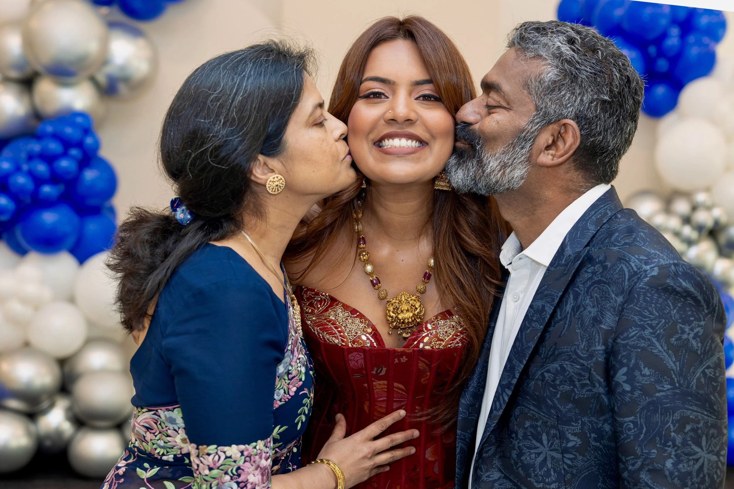 Three people smiling and sharing a moment, two women on either side of a woman in the middle. The woman in the middle is smiling broadly, while the other two are kissing her on each cheek. They are dressed in formal attire with colorful patterns and accessories, and there are blue and white balloons in the background, indicating a celebration or special event.