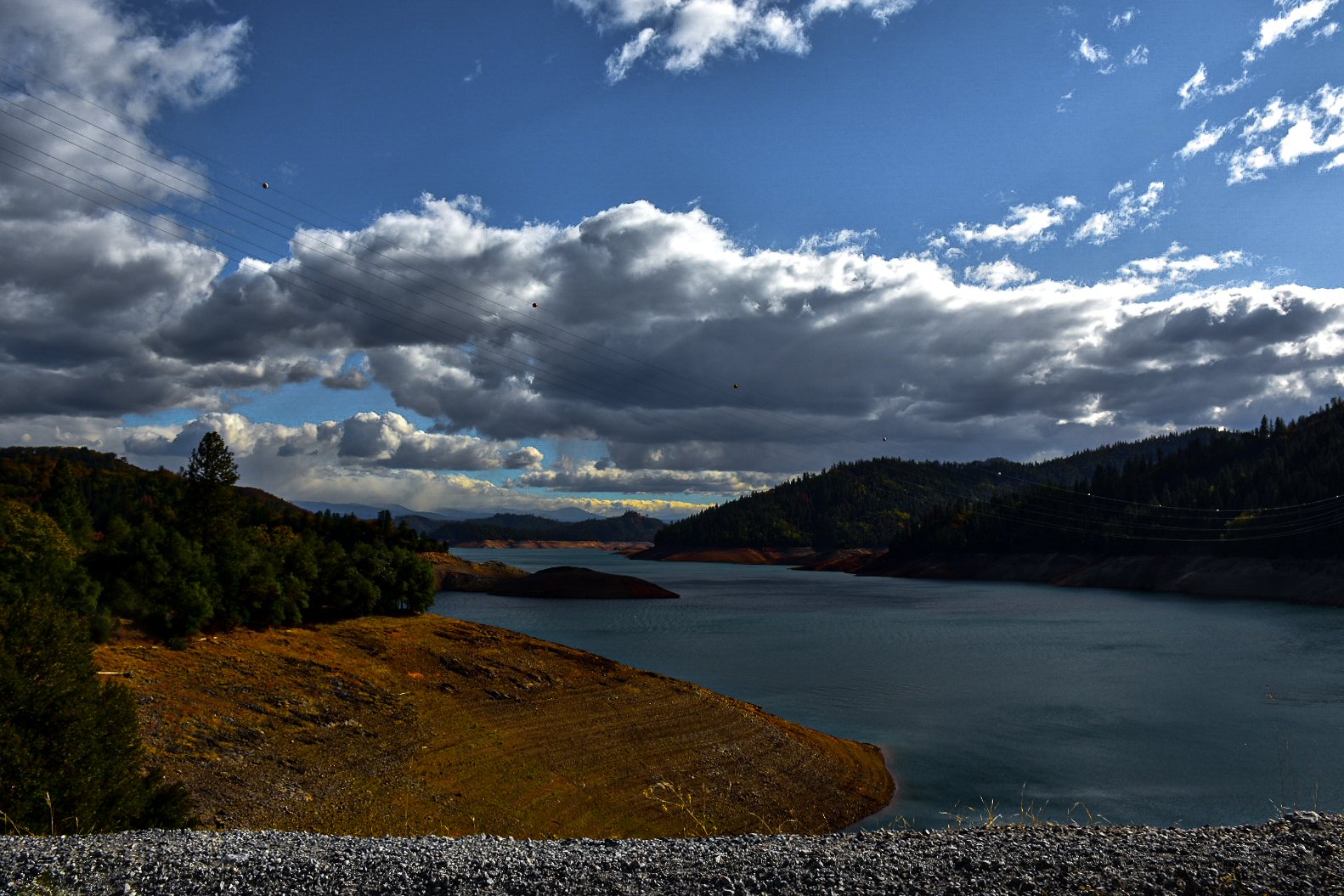 A landscape with a large body of water, surrounded by green hills and mountains under a partly cloudy sky with some power lines crossing.