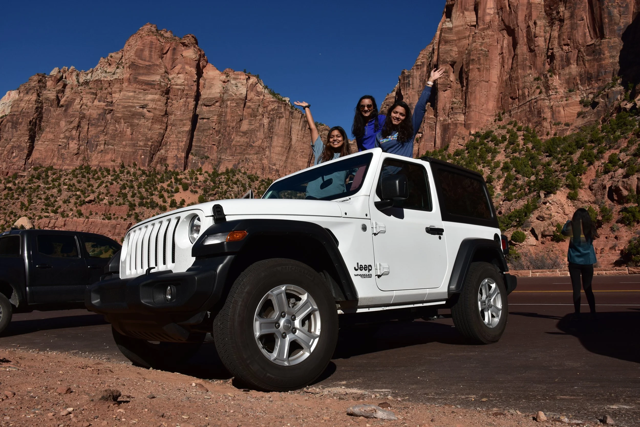 Three women in the back of a white Jeep Wrangler with red rock cliffs in the background, celebrating and posing with their arms raised on a bright sunny day.