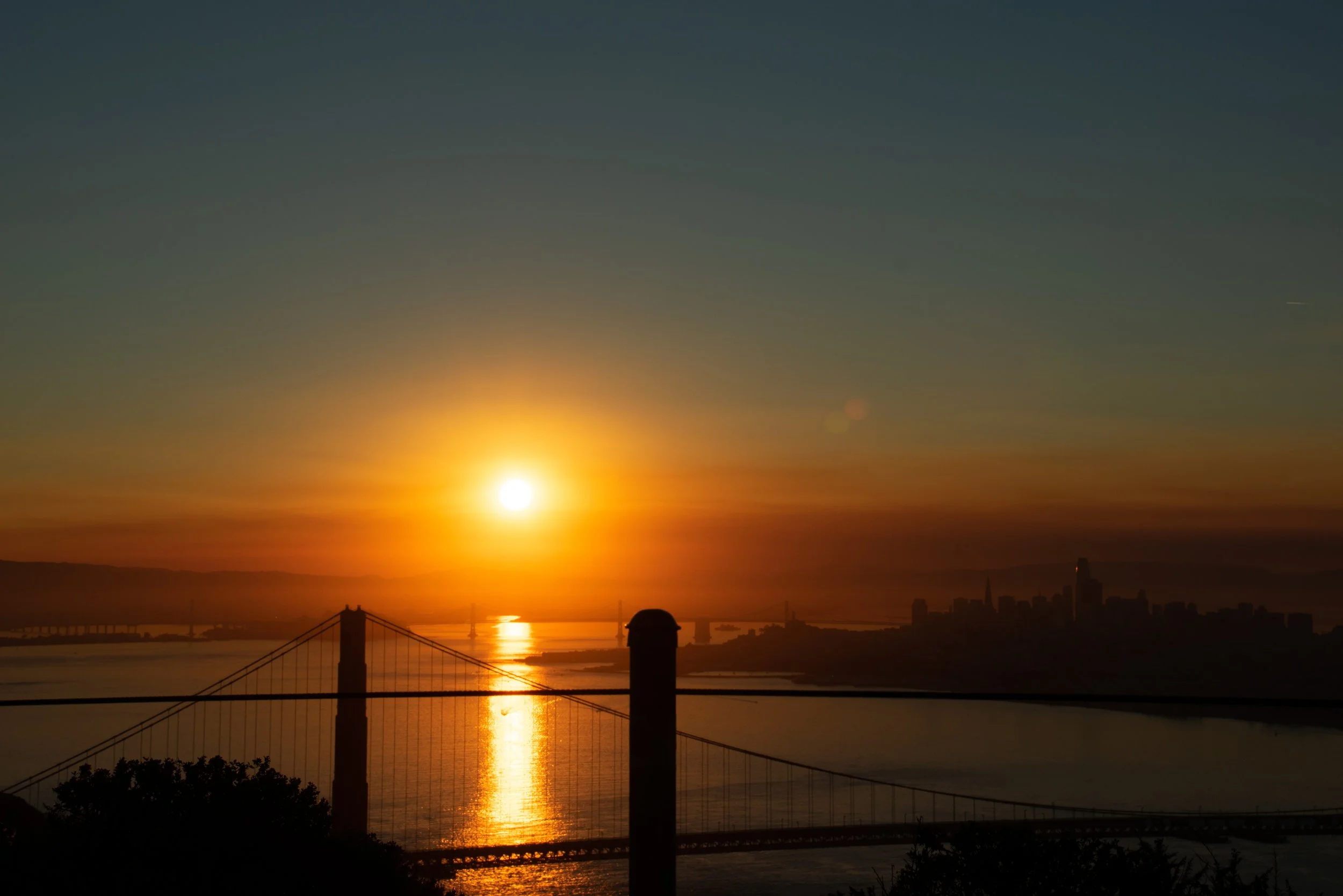 Sunset over a city skyline with a bridge and water reflecting the sunlight.