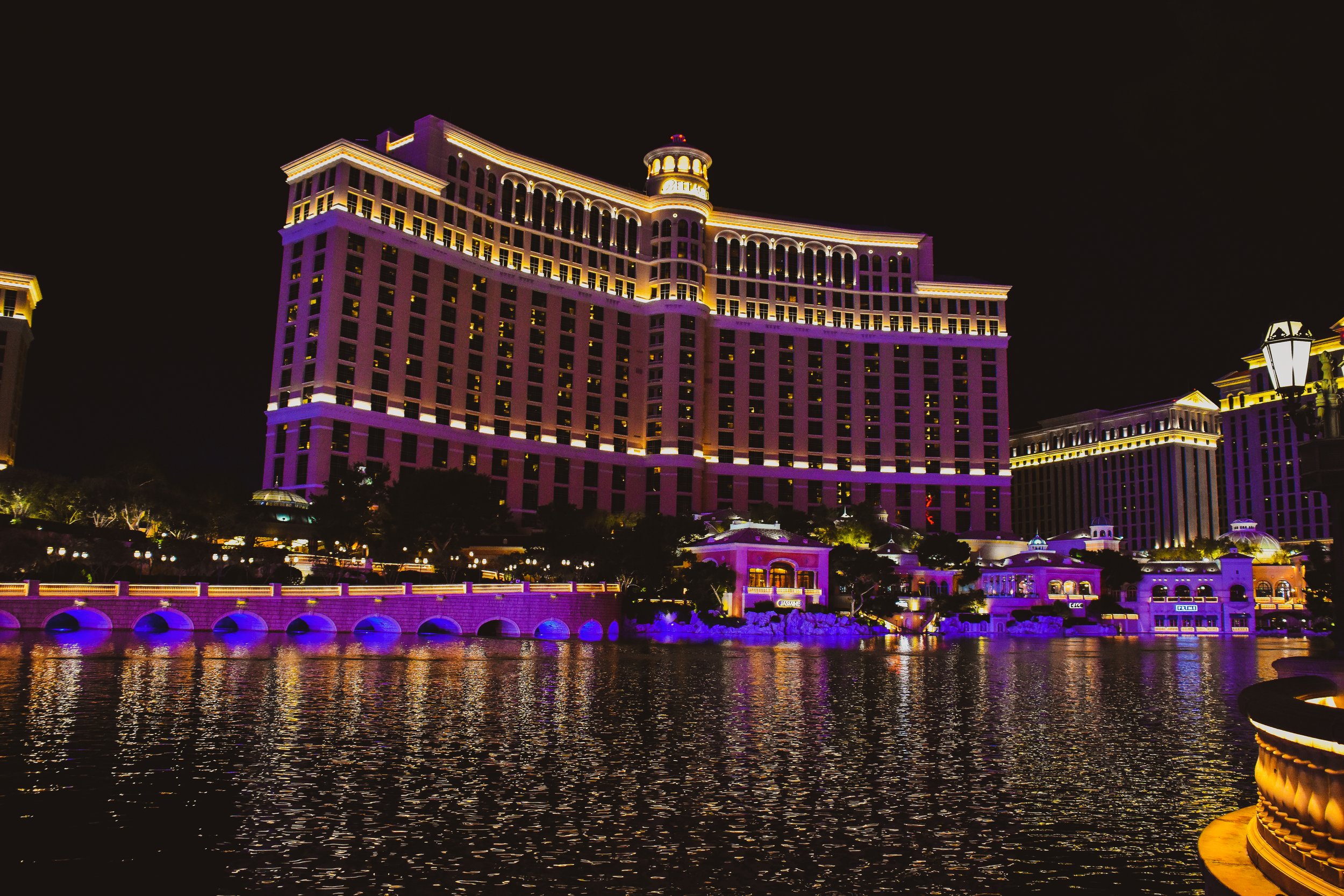 Night view of a brightly lit hotel building with purple and yellow lighting reflections on the water in front.