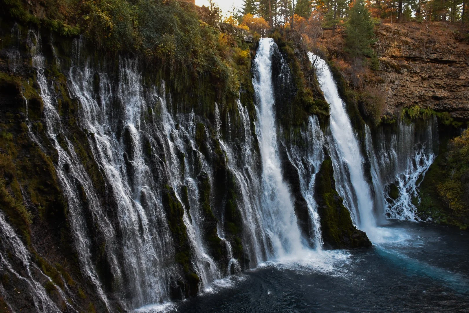 Multiple waterfalls cascading down a rocky cliffside surrounded by lush trees with autumn leaves, flowing into a river below.