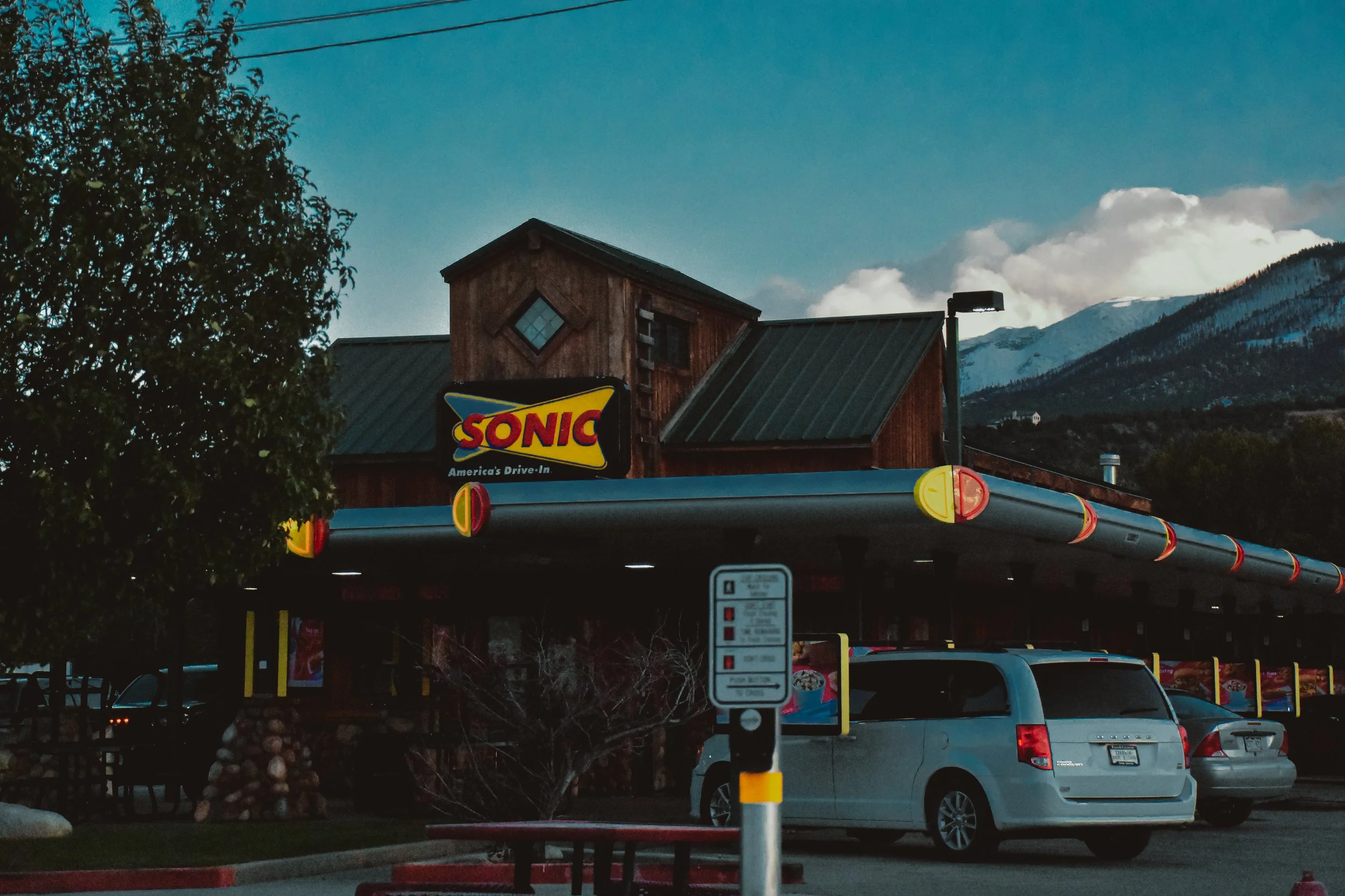 Parking lot with multiple vehicles and a Sonic drive-in restaurant sign, wooden building, mountain backdrop, and cloudy sky.