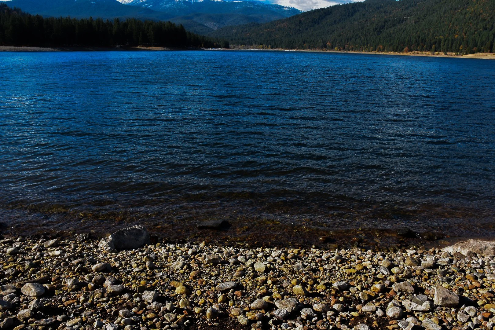 View of a mountain lake with clear blue water, surrounded by dense pine forests and mountains in the background, with a rocky shoreline in the foreground.