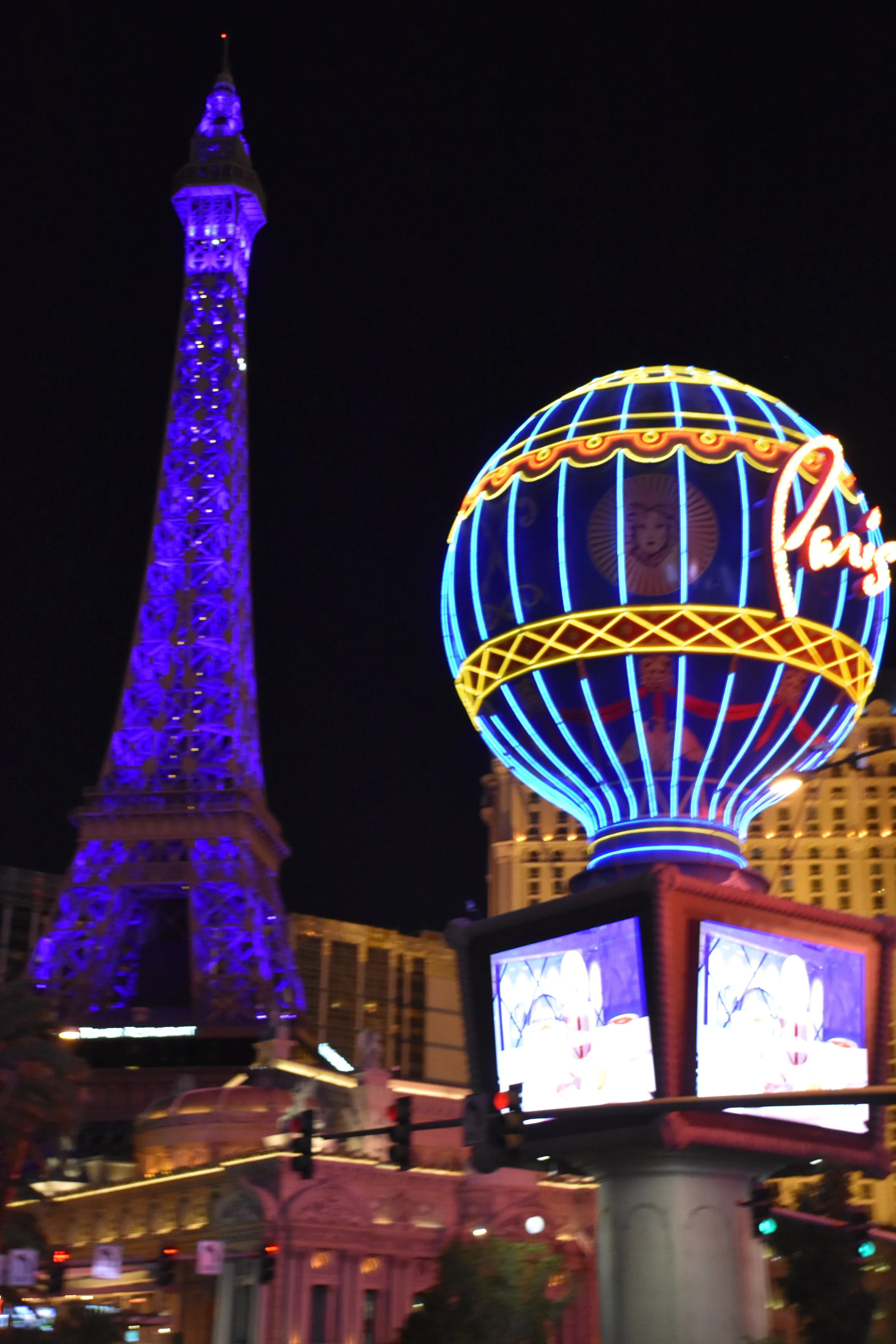 Nighttime view of the Las Vegas Strip with the Paris Las Vegas Hotel's Eiffel Tower replica illuminated in blue and purple lights, and the Parisian-themed hot air balloon sign glowing with neon lights.