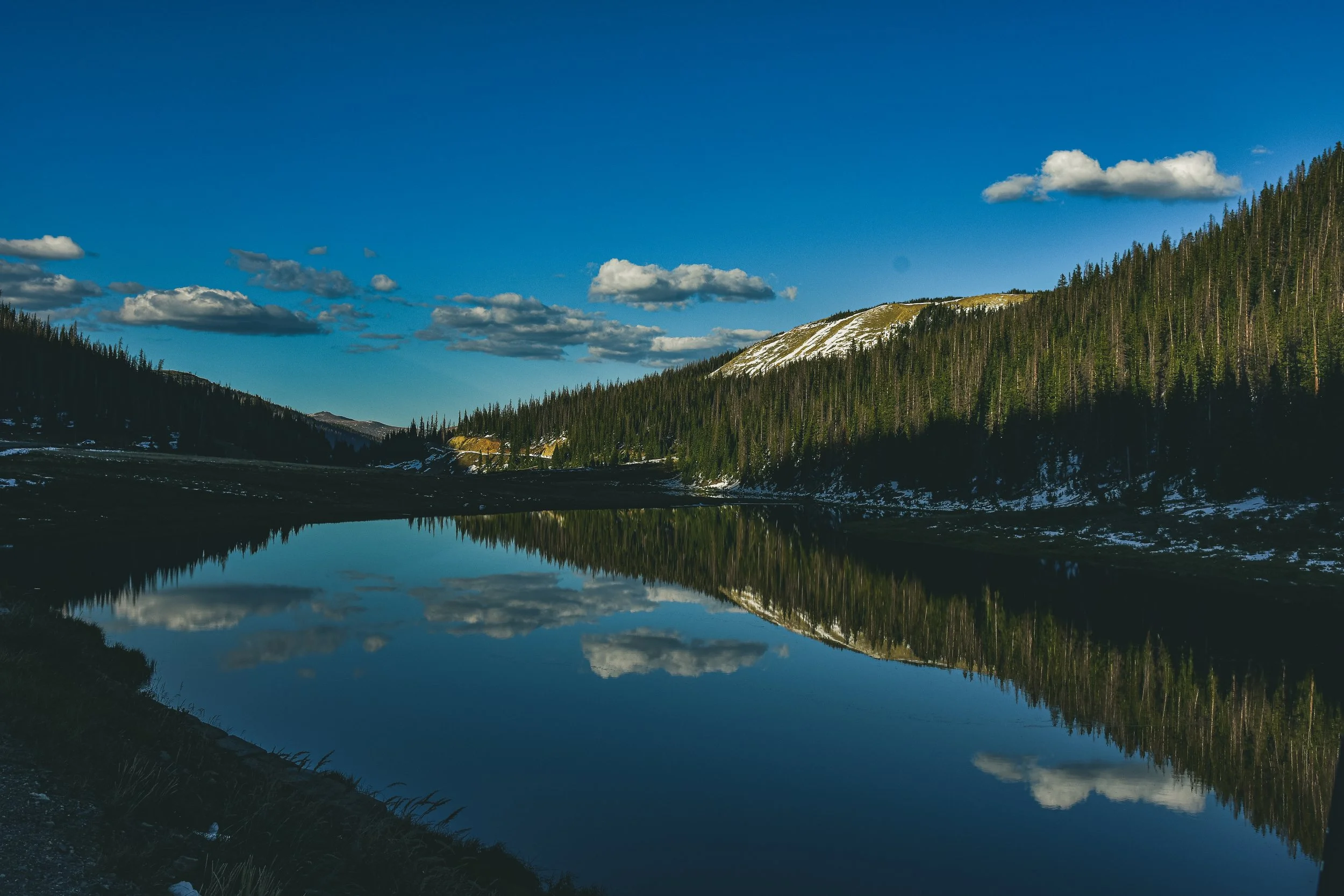 A landscape with a calm river reflecting the sky, surrounded by dense green pine trees on the hills and a snowy mountain in the background under a mostly blue sky with some scattered clouds.