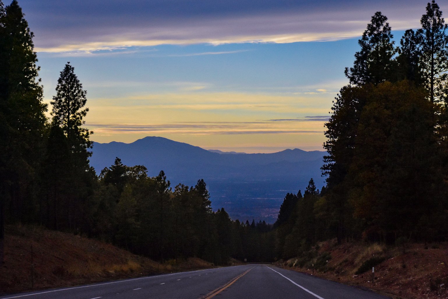 A scenic view of a mountain range in the distance, framed by tall evergreen trees on either side of a winding paved road at sunset, with colorful sky featuring layers of clouds.