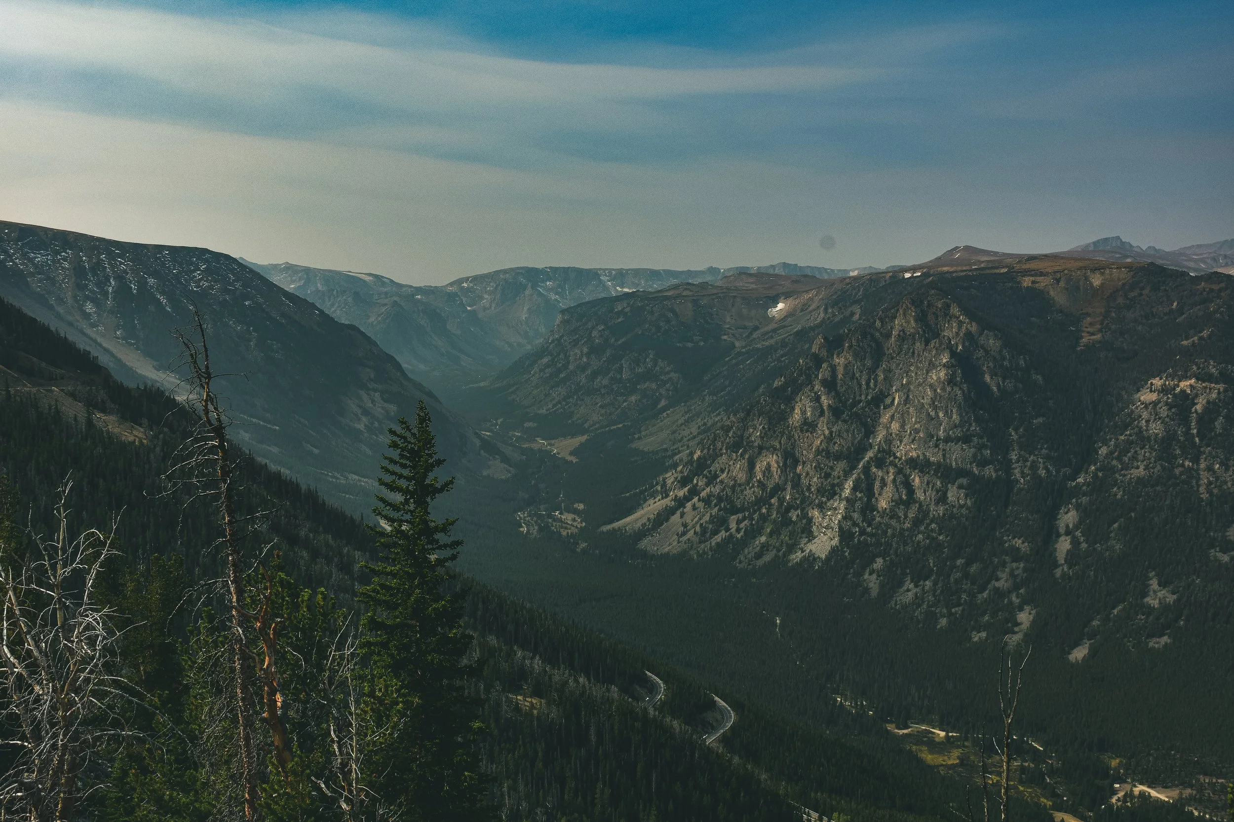 Mountain landscape with steep rocky slopes, green forests, and a winding road below, under a partly cloudy sky.