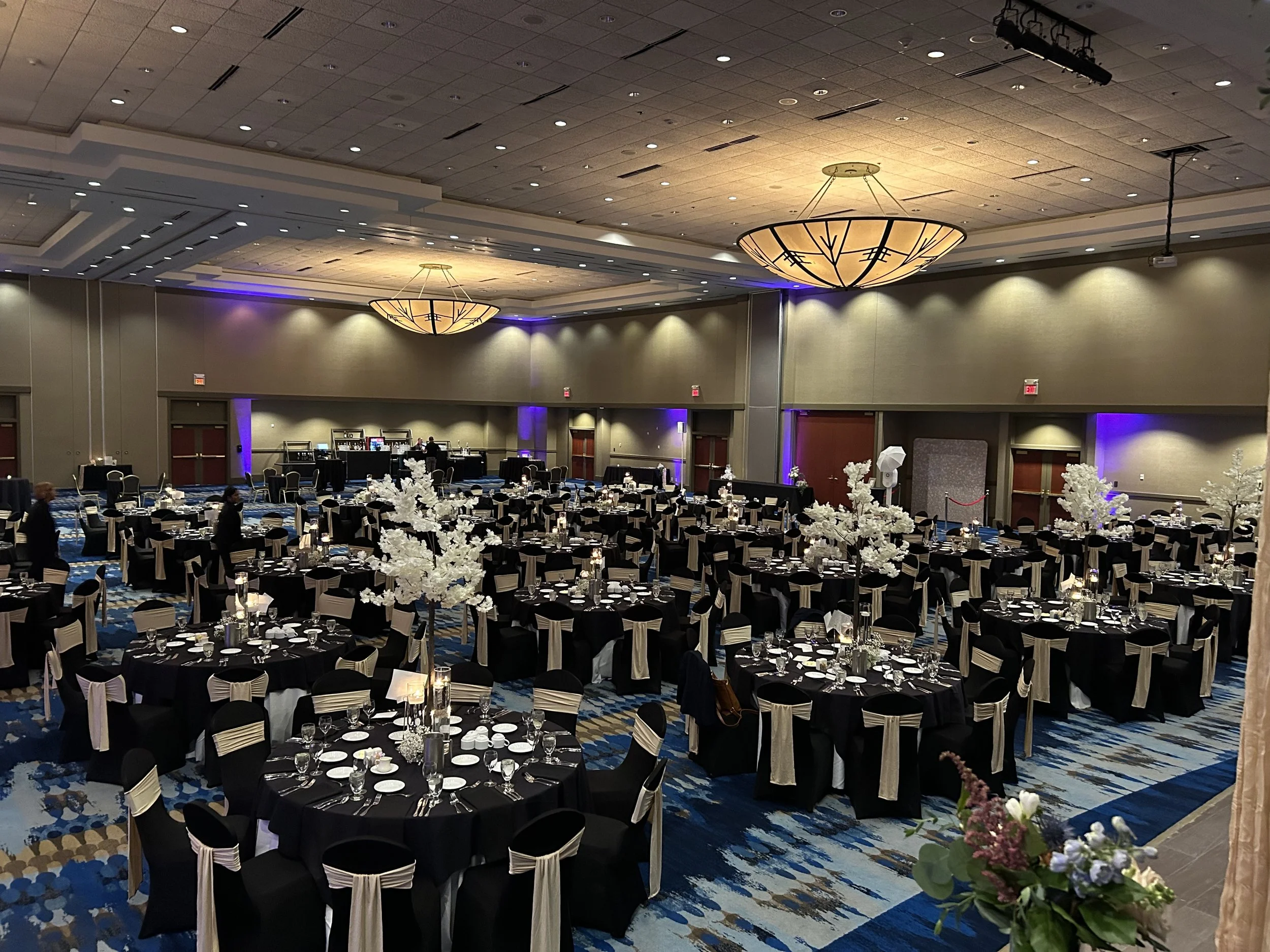 Large banquet hall decorated for an event with round tables covered in black tablecloths, white chair sashes, tall white floral centerpieces, and candles. Dim lighting with purple accents, high ceiling with large chandeliers, and a blue patterned carpet.