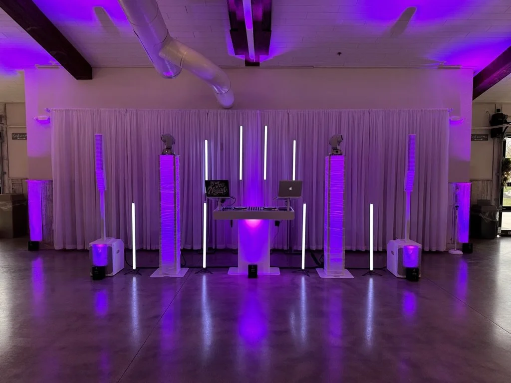 DJ setup with purple lighting, including speakers, lighting columns, laptop on a stand, and decorative curtains in the background. Event venue with industrial ceiling design.