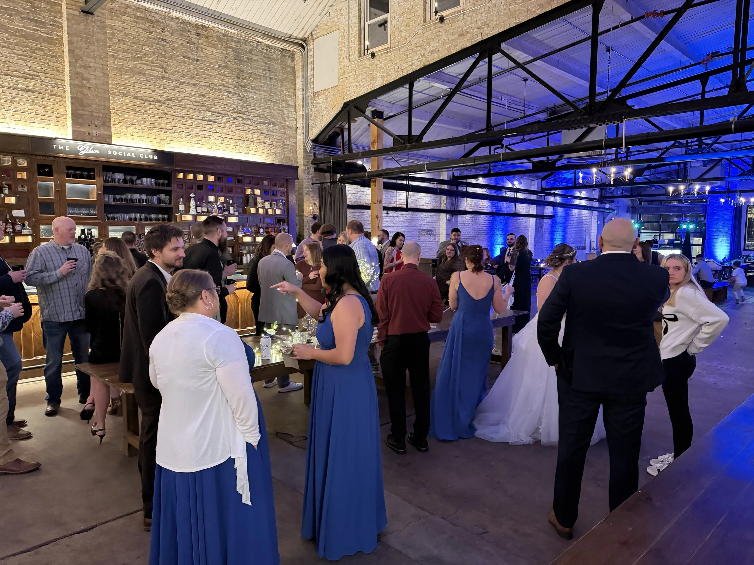 People socializing at a party in a spacious industrial-style venue with a bar, brick walls, and blue lighting.