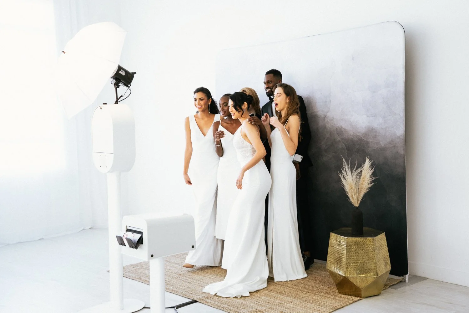 Group of six diverse people dressed in white and black formal attire posing for a photo in a photo studio with a black backdrop, standing on a tan rug next to a gold decorative planter with dried pampas grass.