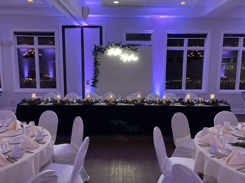 Elegant banquet hall setup with round tables, white tablecloths, and chairs. Long table at the back with a greenery garland and candles, under a glowing sign. Purple uplighting enhances the ambience.