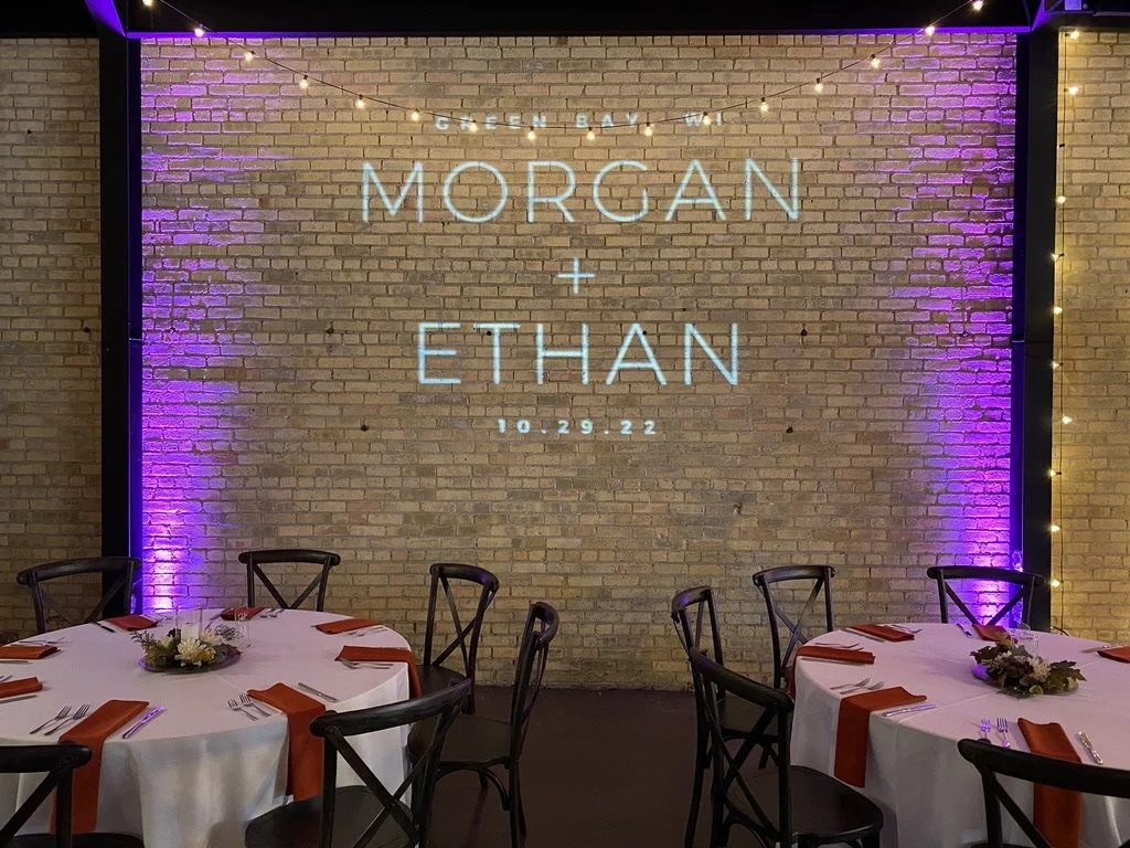 Wedding venue setup with round tables, white tablecloths, orange napkins, floral centerpieces, and a brick wall with string lights, featuring a projection of wedding details: "Morgan + Ethan 10.29.22" in Green Bay, WI.