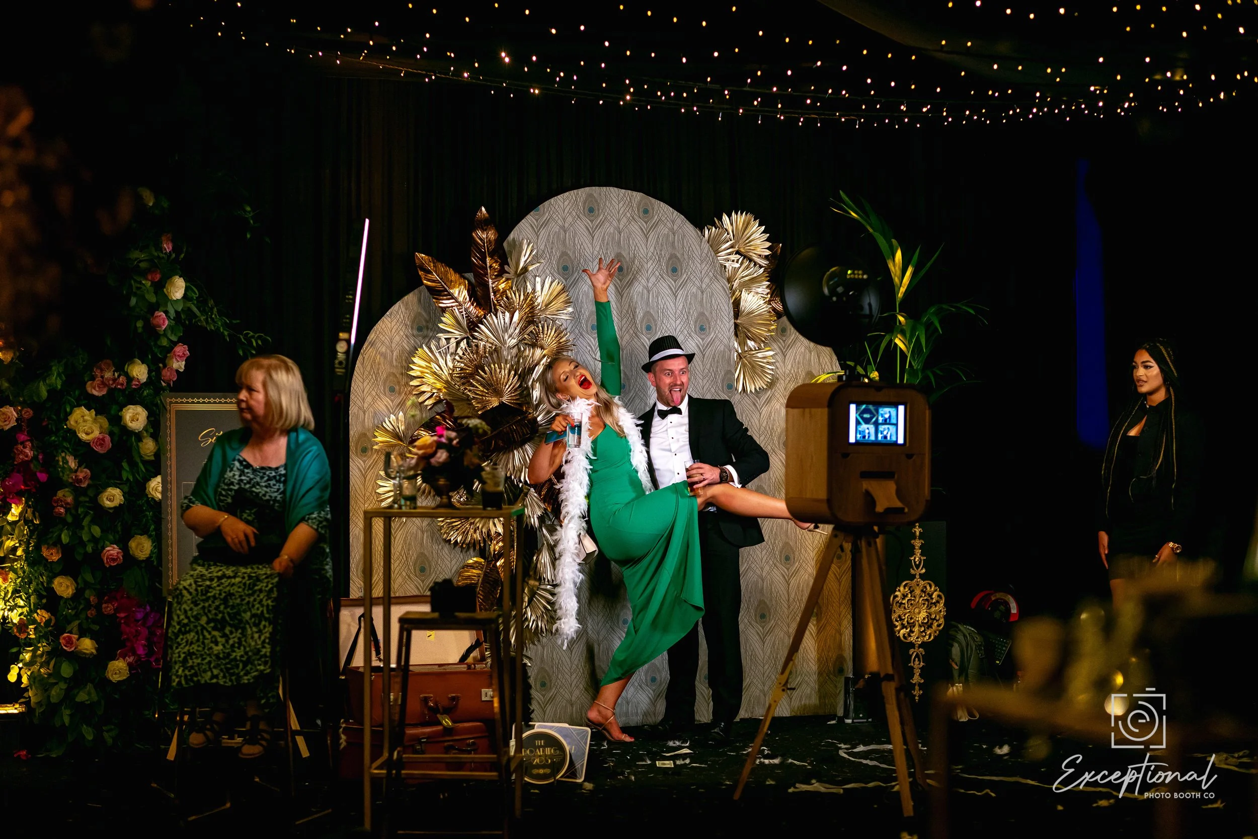 People celebrating at a festive event with a decorated backdrop, including a woman in a green dress and hat, holding a drink and posing playfully with a man in a tuxedo, album covers and gold decorations around, and a camera on tripod capturing the moment.