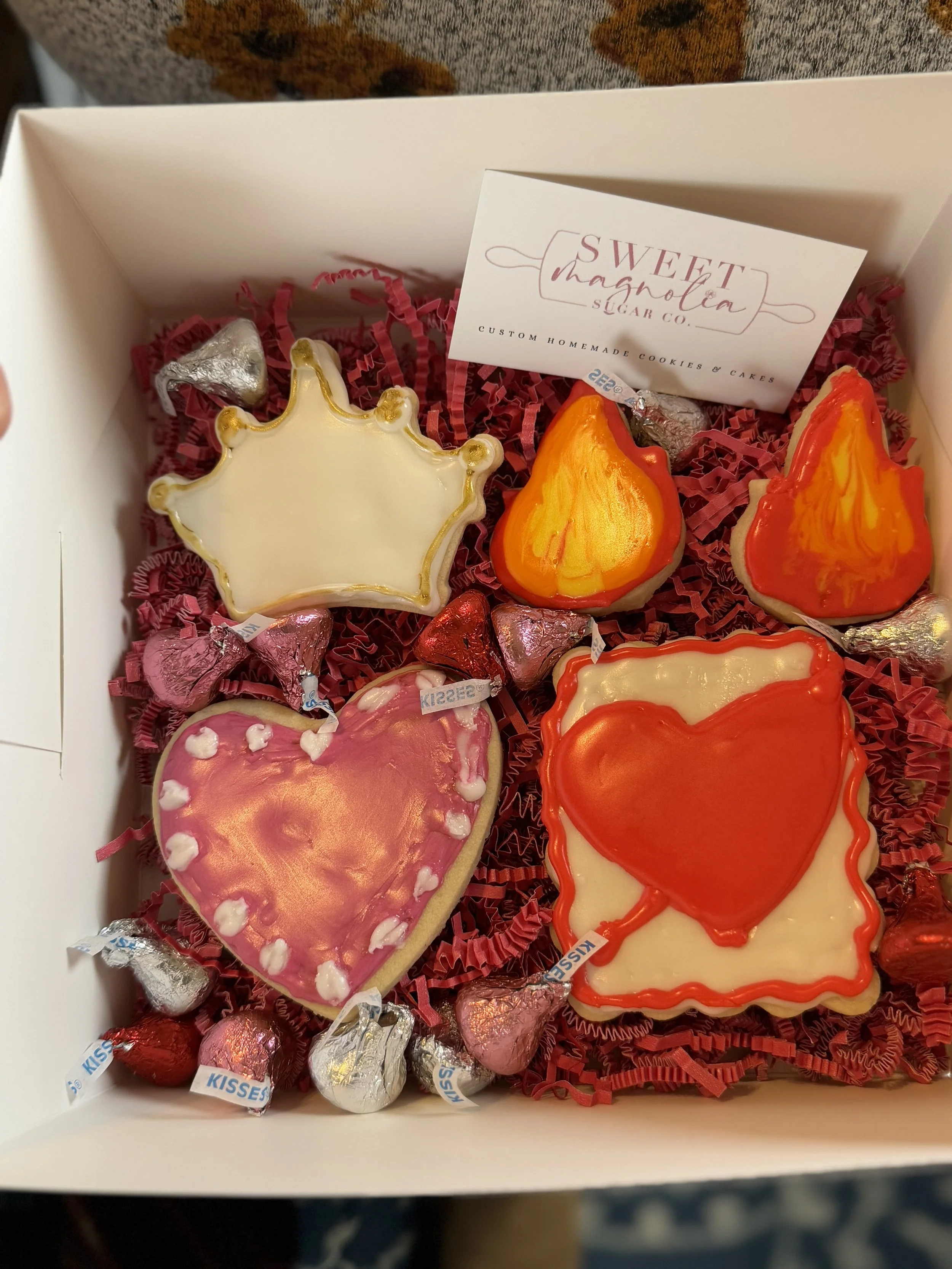 Box of decorated cookies with pink and red hearts, a crown, and heart-shaped cookies with Valentine-themed icing, surrounded by metallic-wrapped chocolates.