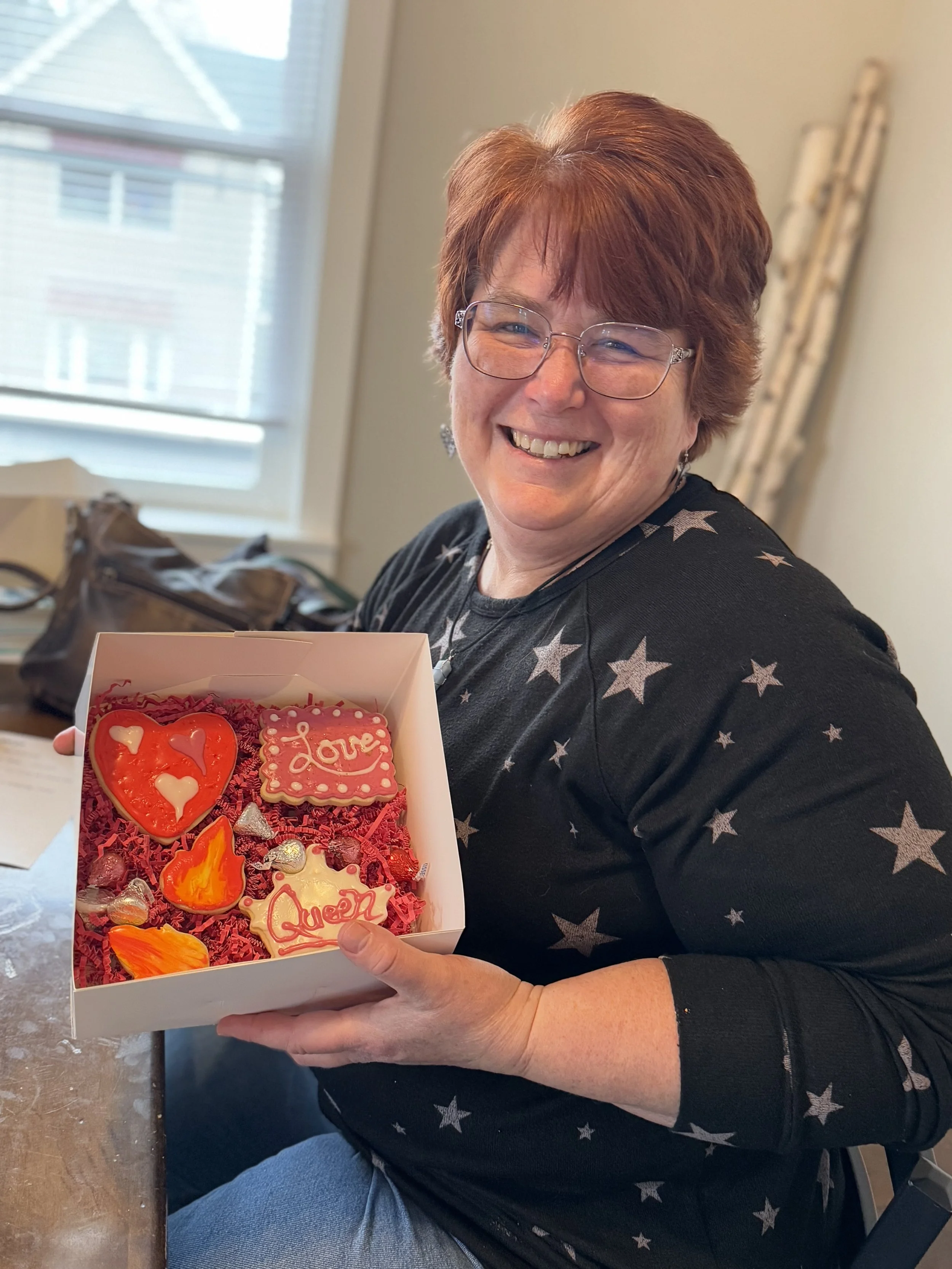 A woman with red hair and glasses smiling while holding a box of Valentine's cookies decorated with hearts and 'love' messages.