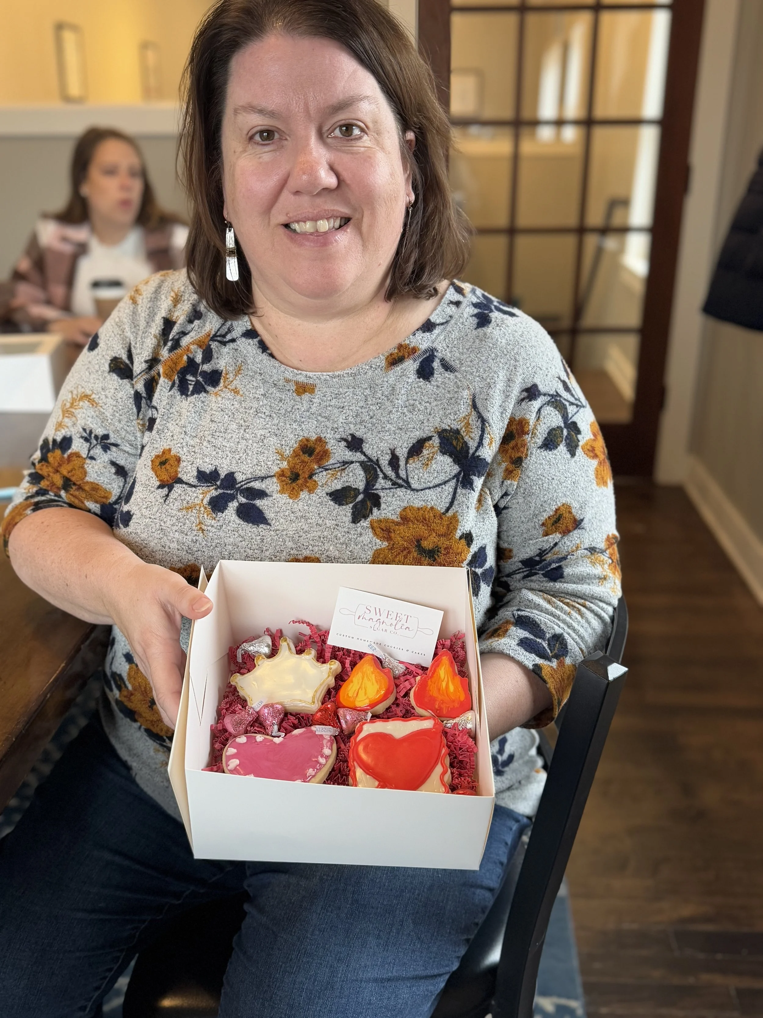 A woman smiling and holding a box of decorated cookies shaped like hearts and flames.