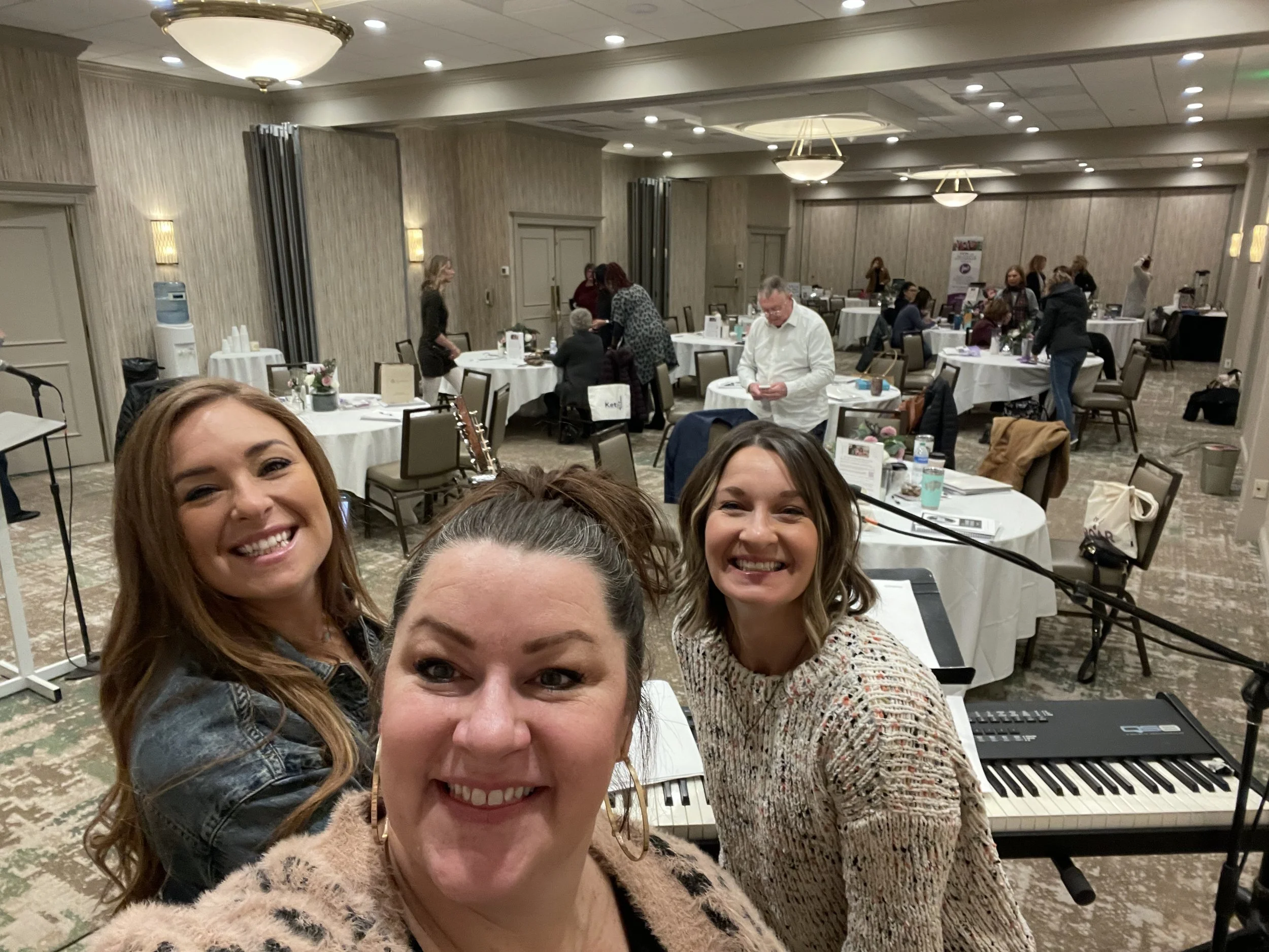 Three women smiling and taking a selfie at a conference or banquet hall with round tables, chairs, and people in the background.