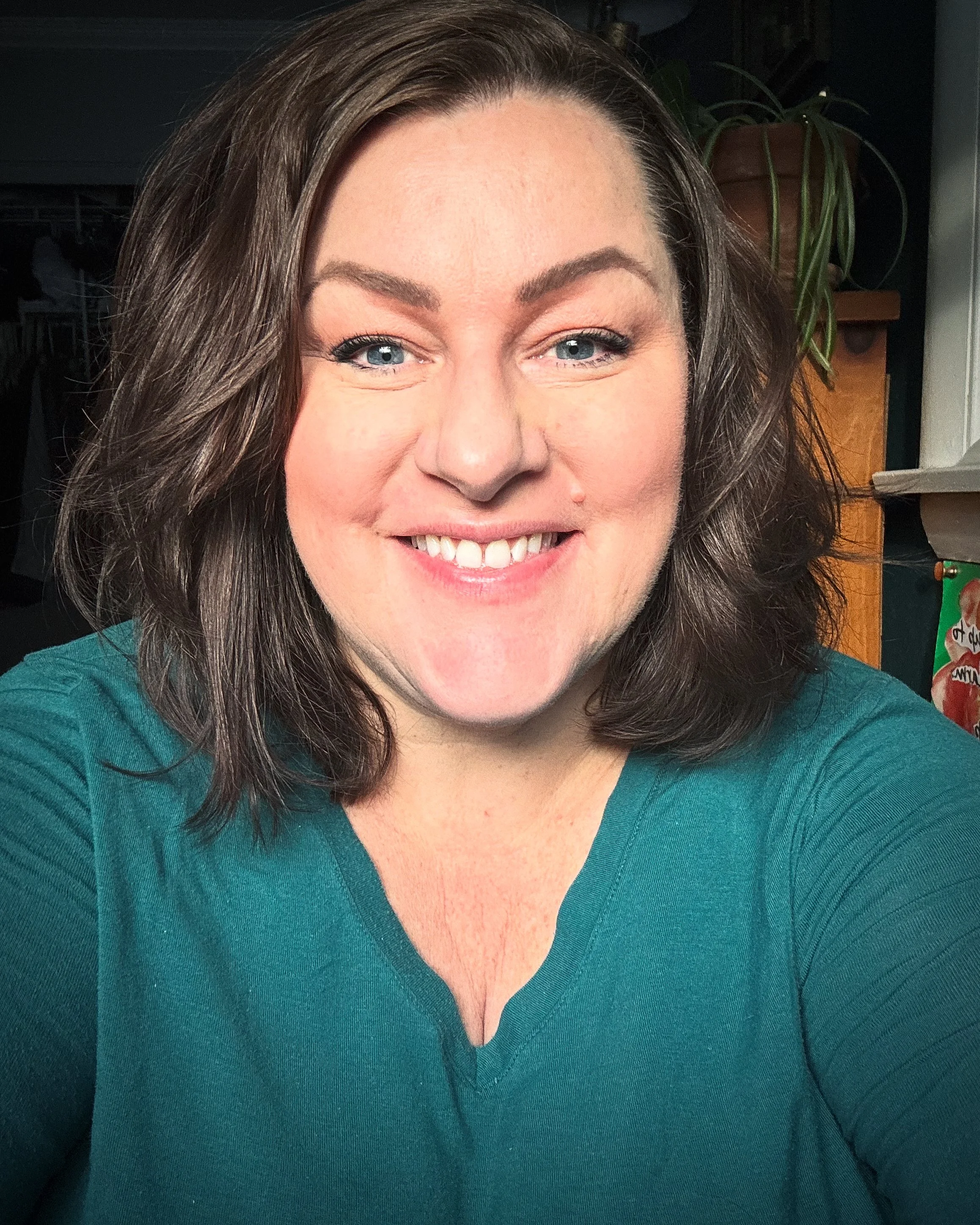 CHRISTINE WELCH 
A smiling woman with shoulder-length brown hair and blue eyes wearing a teal shirt, sitting indoors with a houseplant and some furniture in the background.