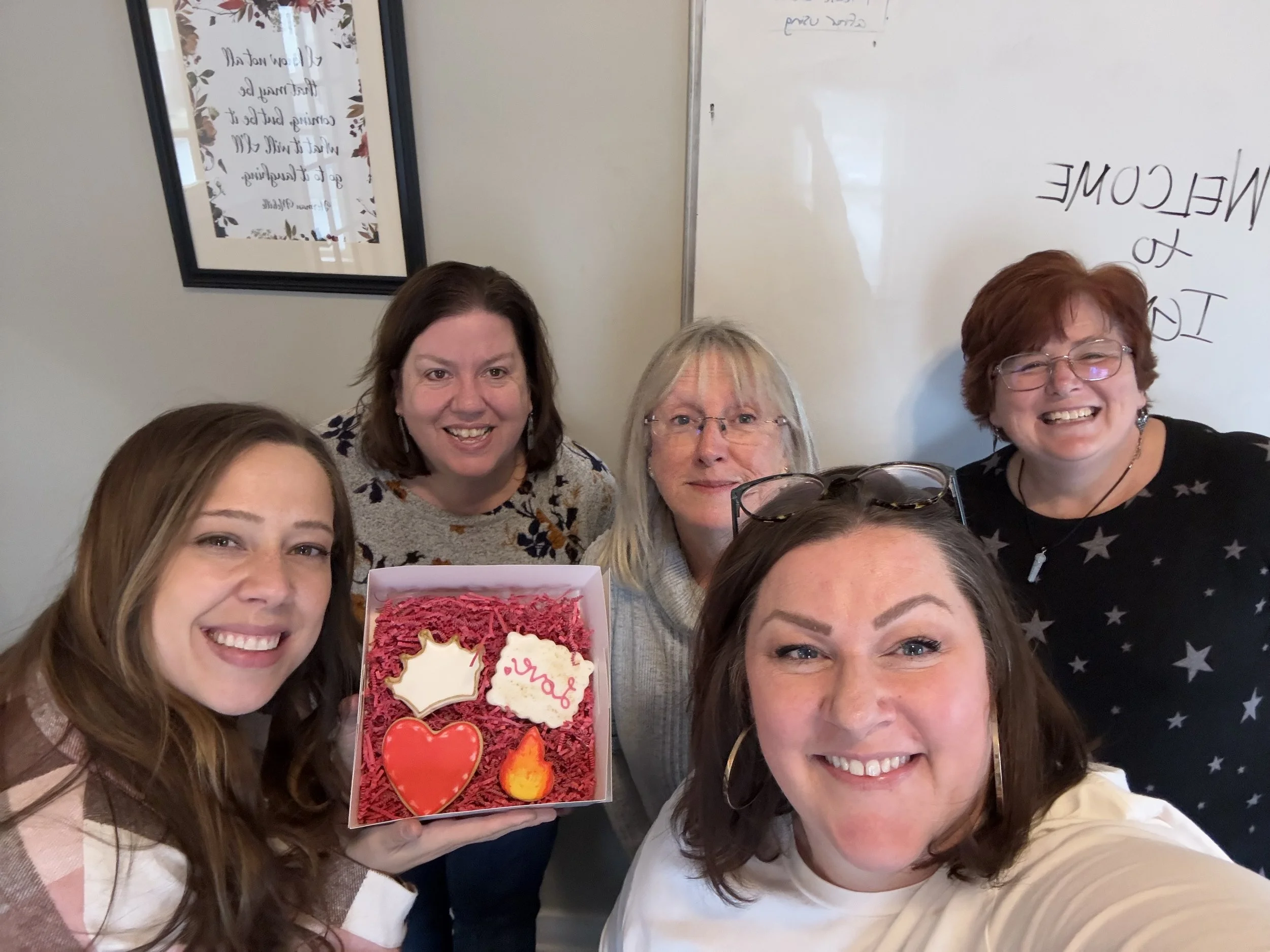 A group of five women smiling, holding a decorated box with various cookies and treats, in an indoor setting with a whiteboard and framed picture in the background.