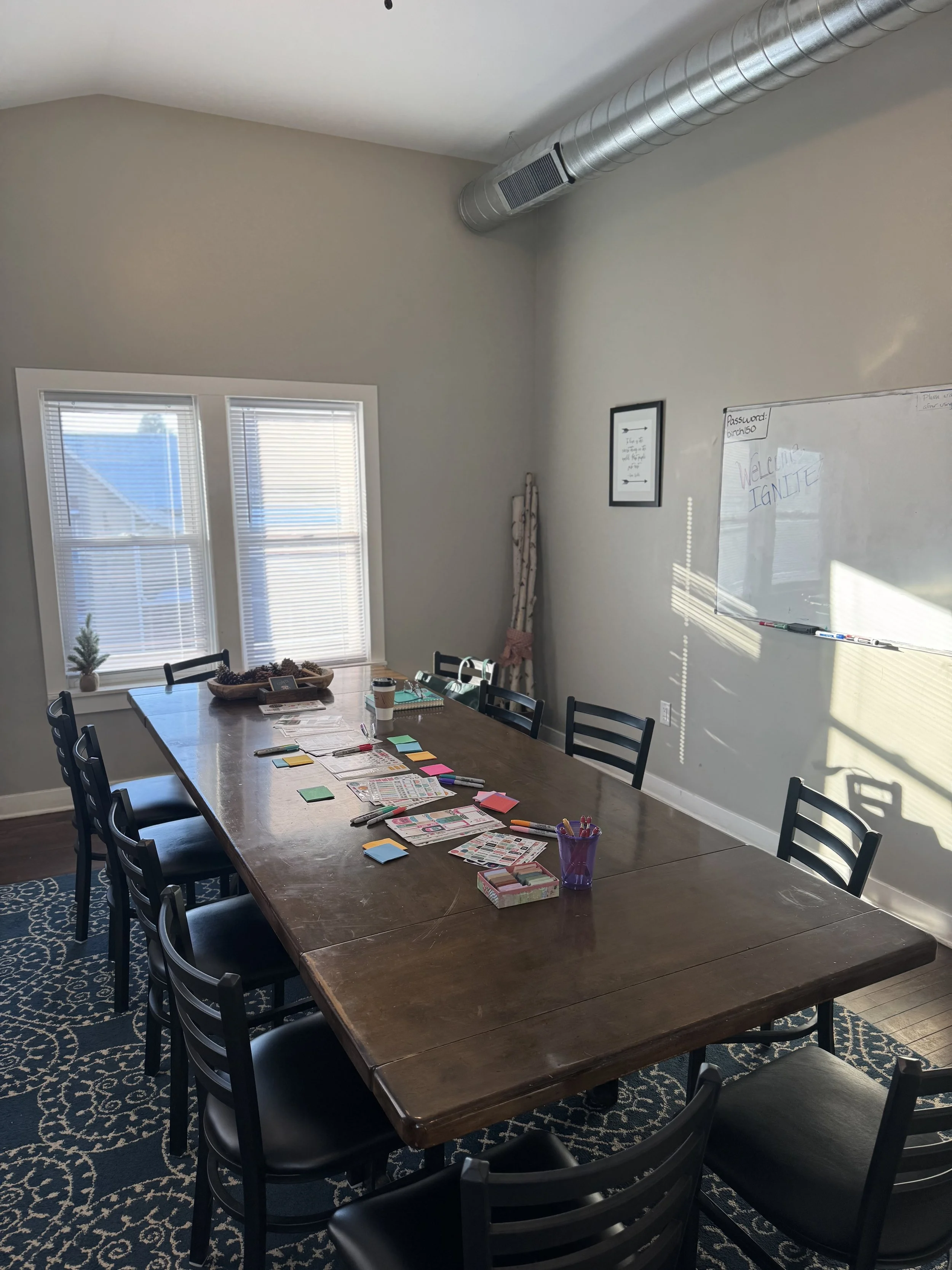 A dining room with a long wooden table surrounded by black chairs, set with colorful sticky notes, markers, and papers, near two windows with blinds and a whiteboard on the wall that reads 'Welcome Ignite'.